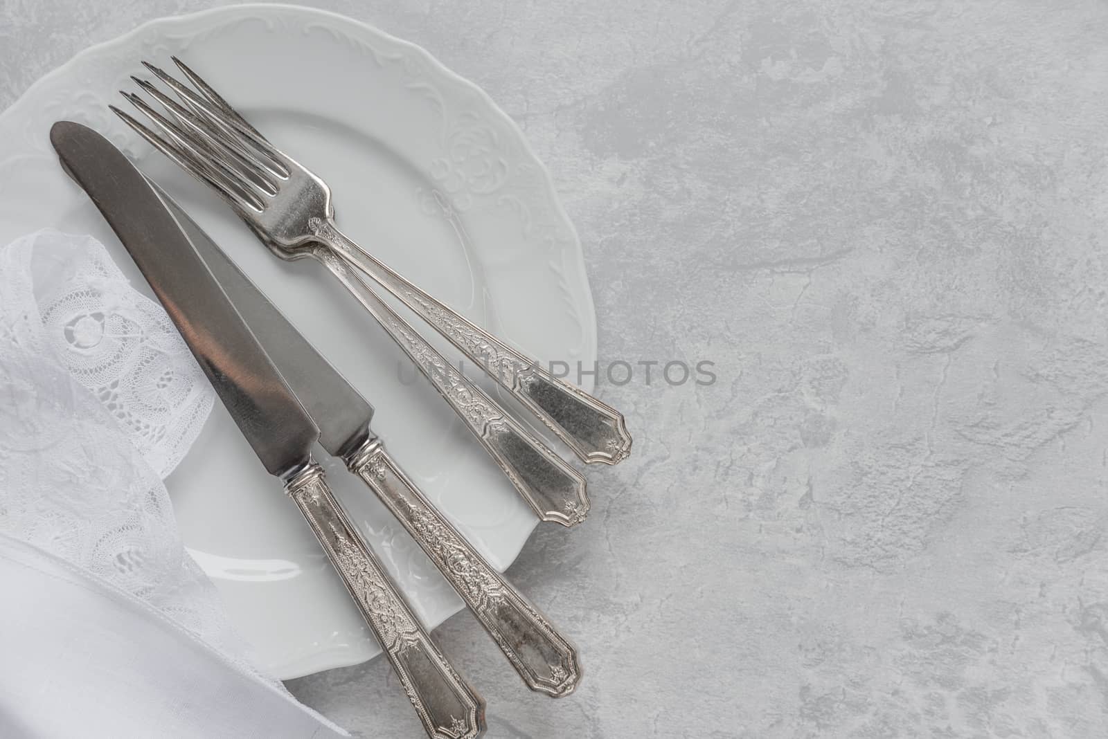 Silver cutlery on a porcelain plate and white napkin with Belgian lace are on the background of gray concrete surface, with copy-space