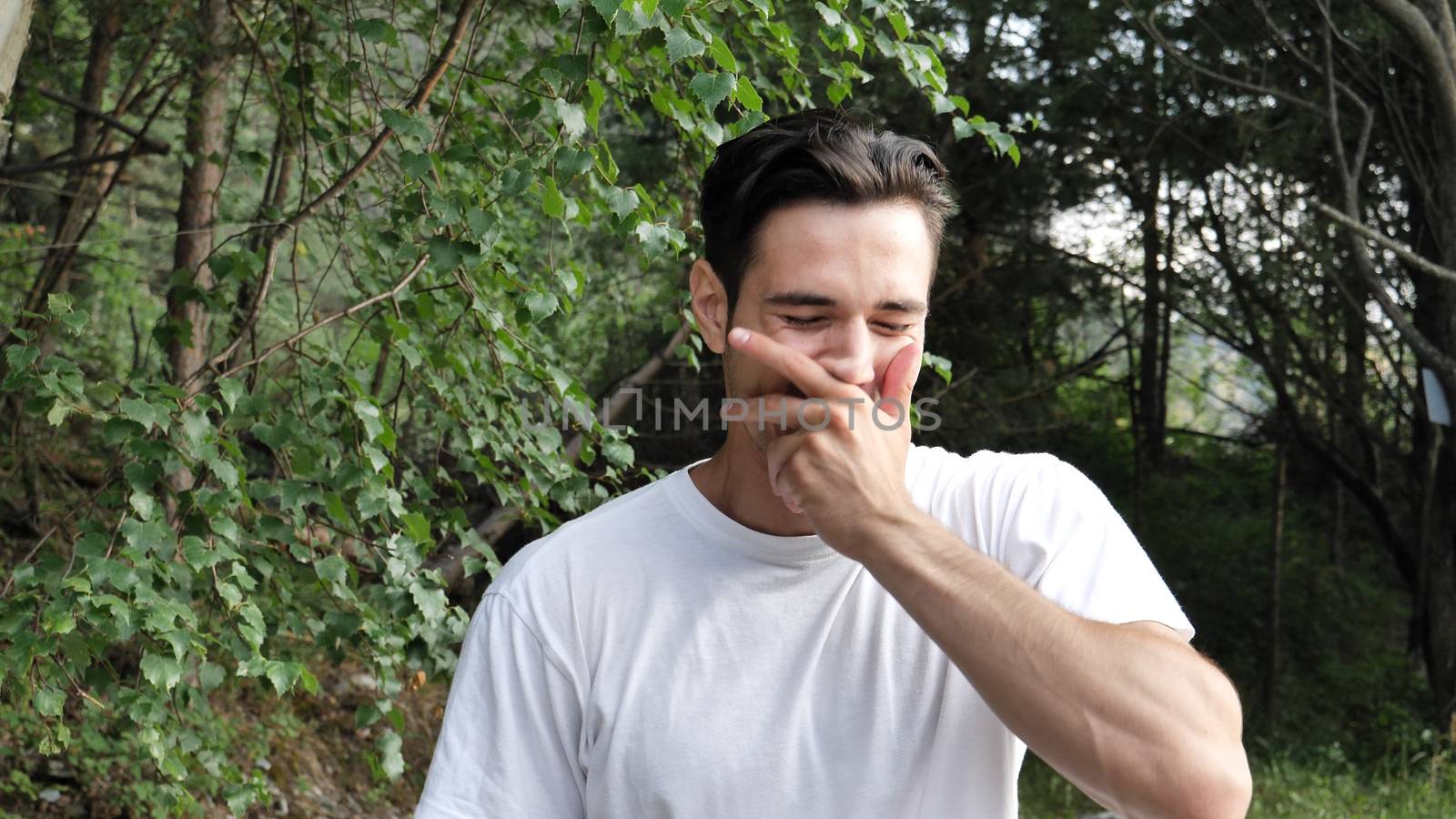 Young handsome man standing in nature, trying not to laugh then bursting into laughter