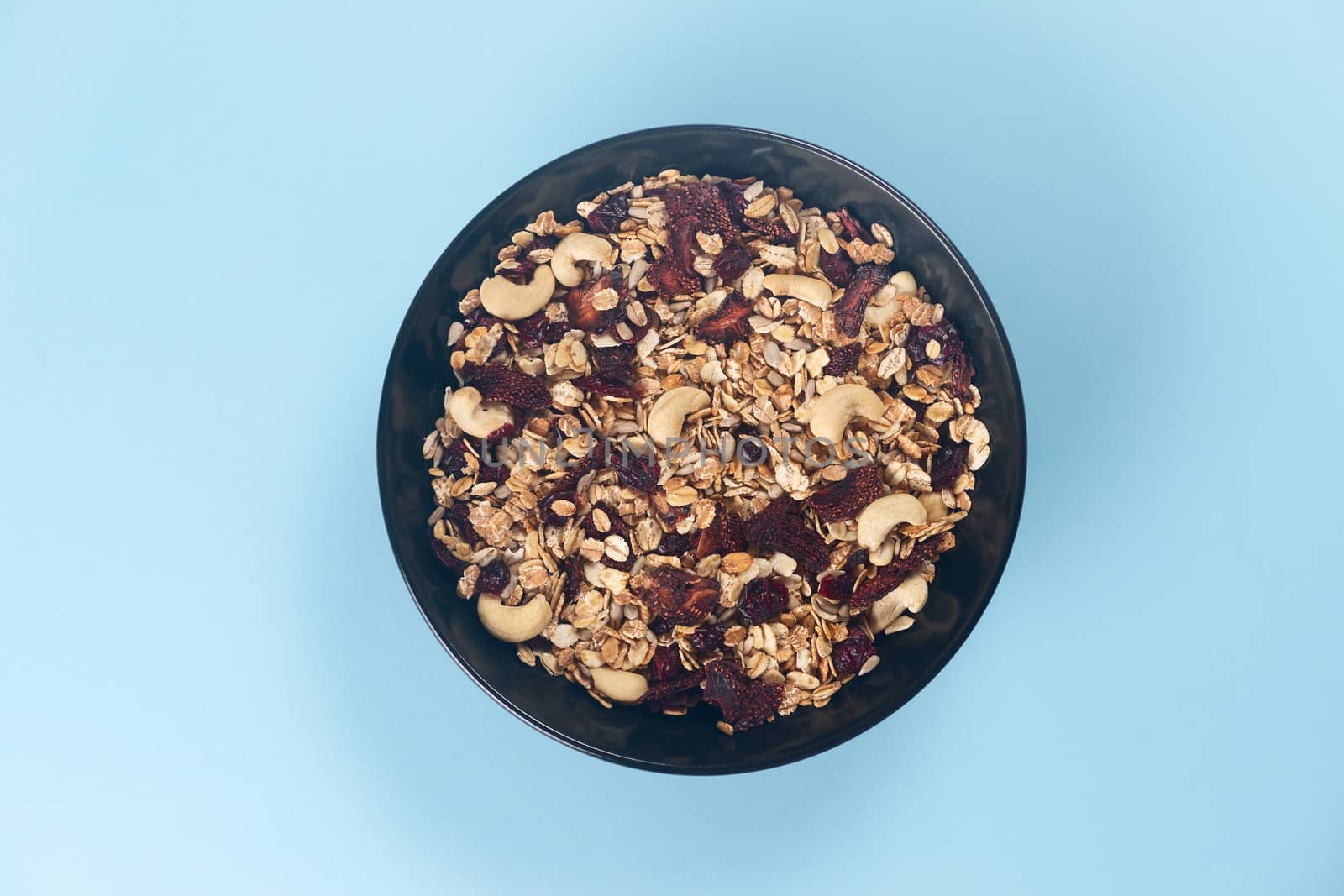 A fresh granola with dried and candied nuts and fruits in black bowl on blue bakground. Concept of nutrient and healty breakfast or meal