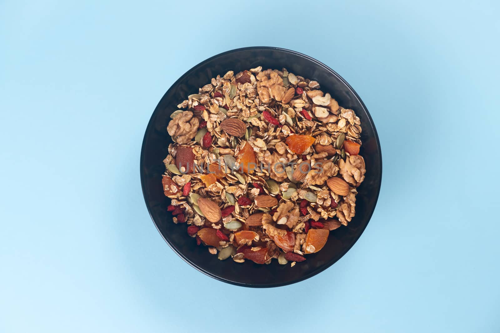 A fresh granola with dried and candied nuts and fruits in black bowl on blue bakground. Concept of nutrient and healty breakfast or meal