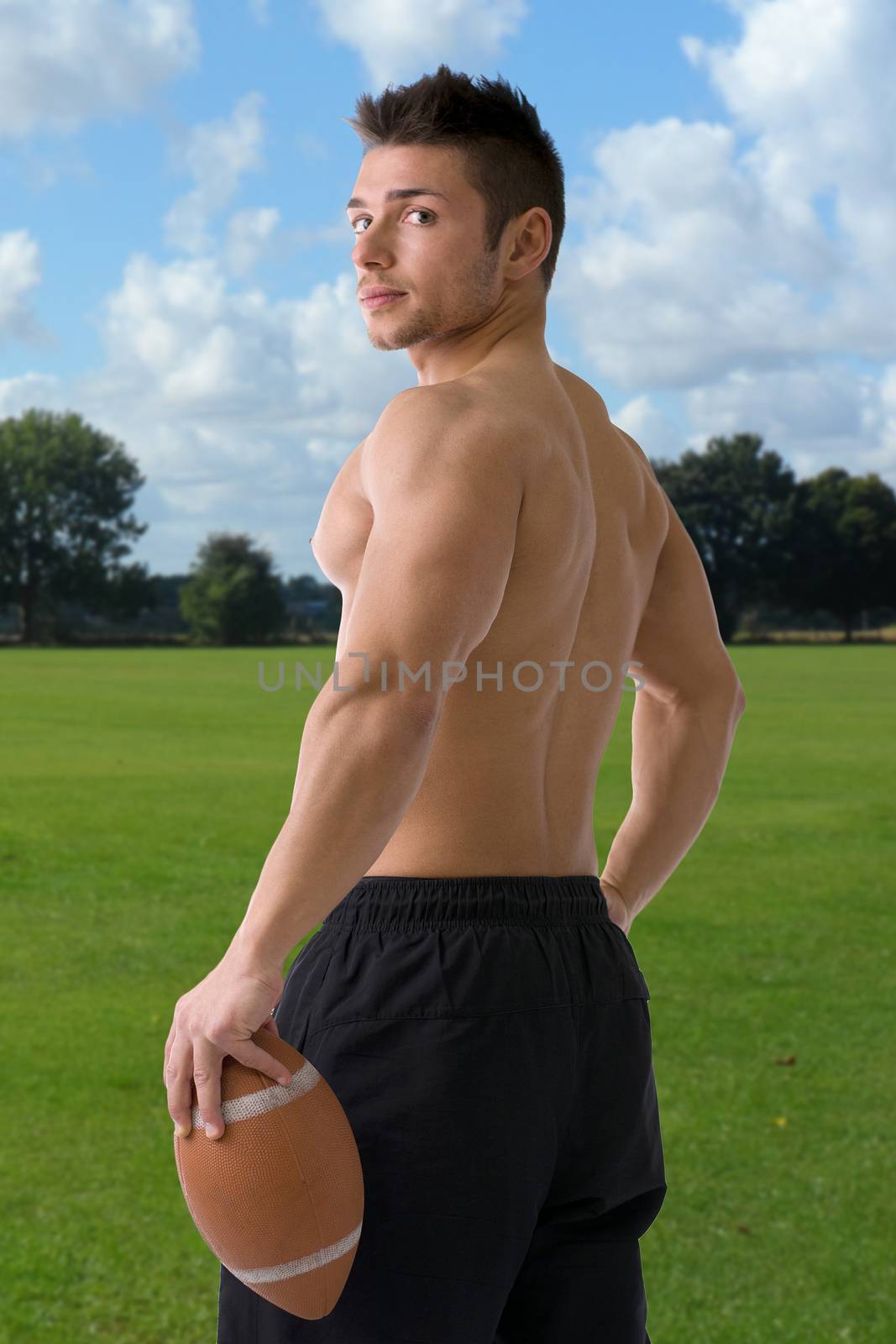 Muscular american football player standing with ball in hand, seen from the back, in playing field