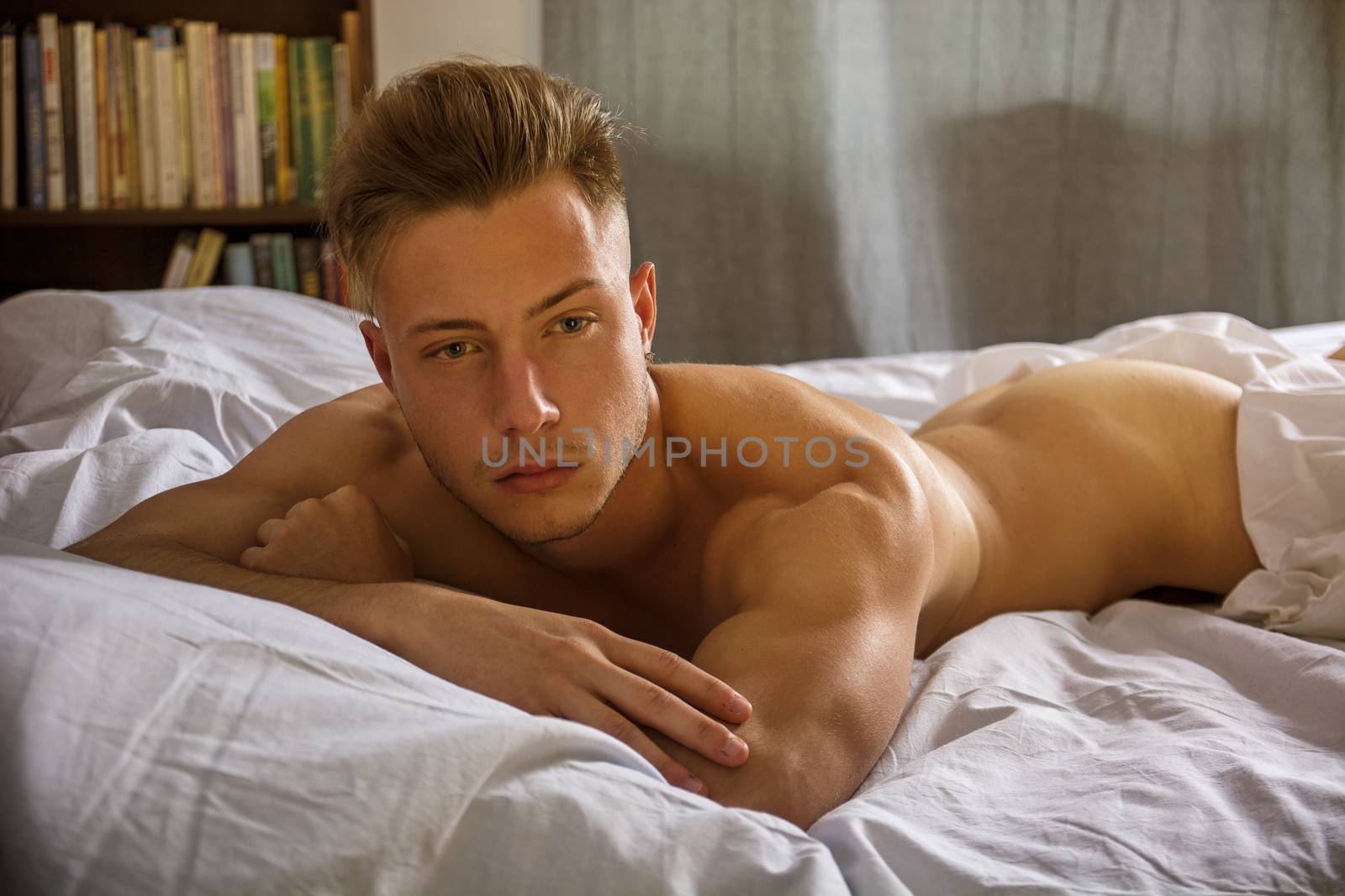 Shirtless sexy male model lying alone on his bed in his bedroom, looking at camera with a seductive attitude