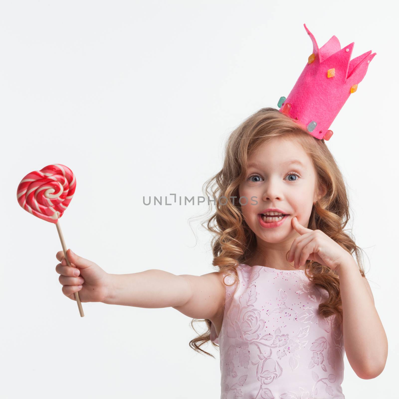 Beautiful little candy princess girl in crown holding big pink heart lollipop and smiling isolated on white