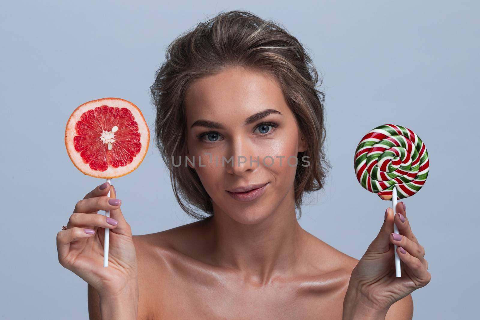 Young woman holding slice of grapefruit and lollipop candy, healthy eating concept, studio shoy on gray background