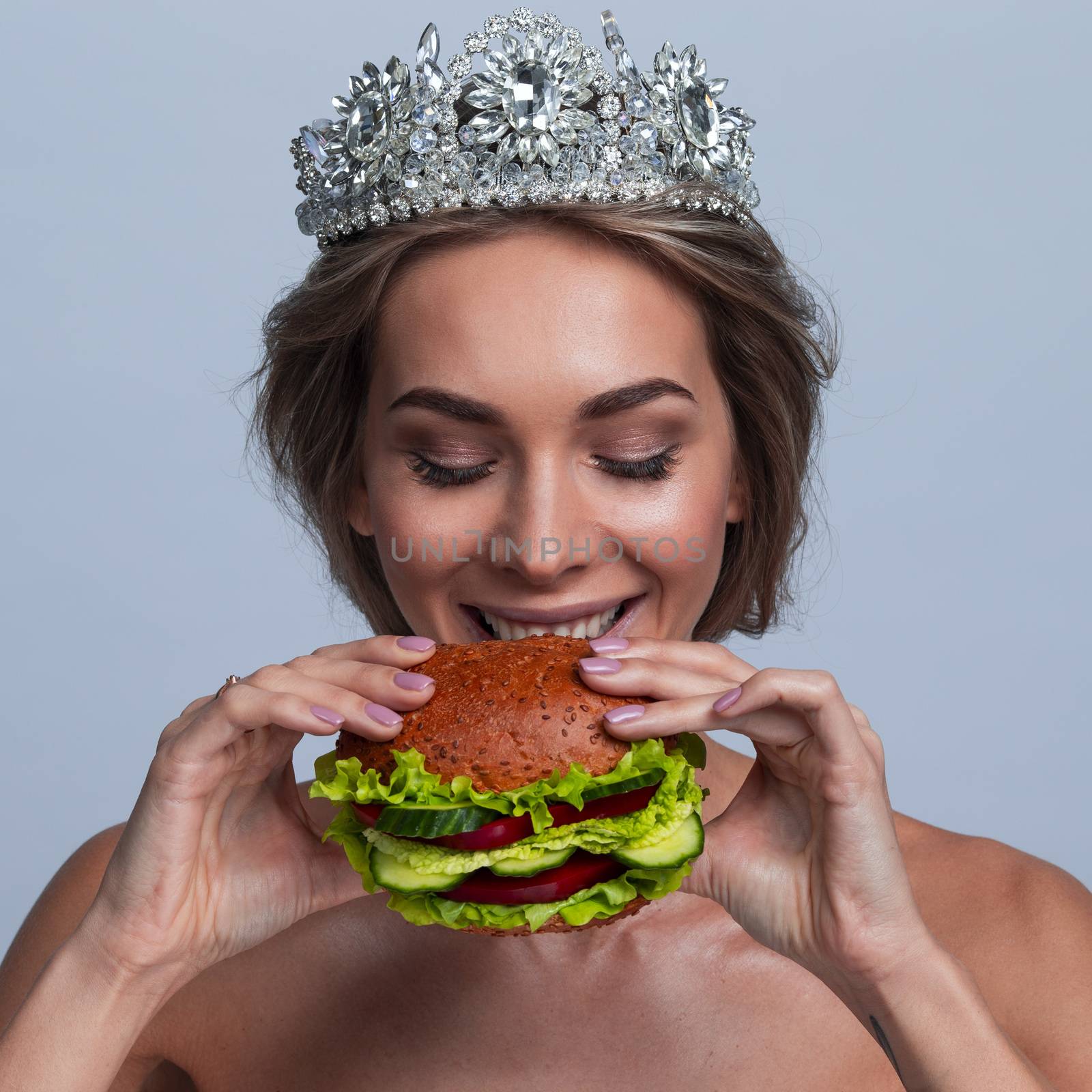 Portrait of beautiful young blonde princess woman with a diadem on a head holding vegan burger made of vegetables