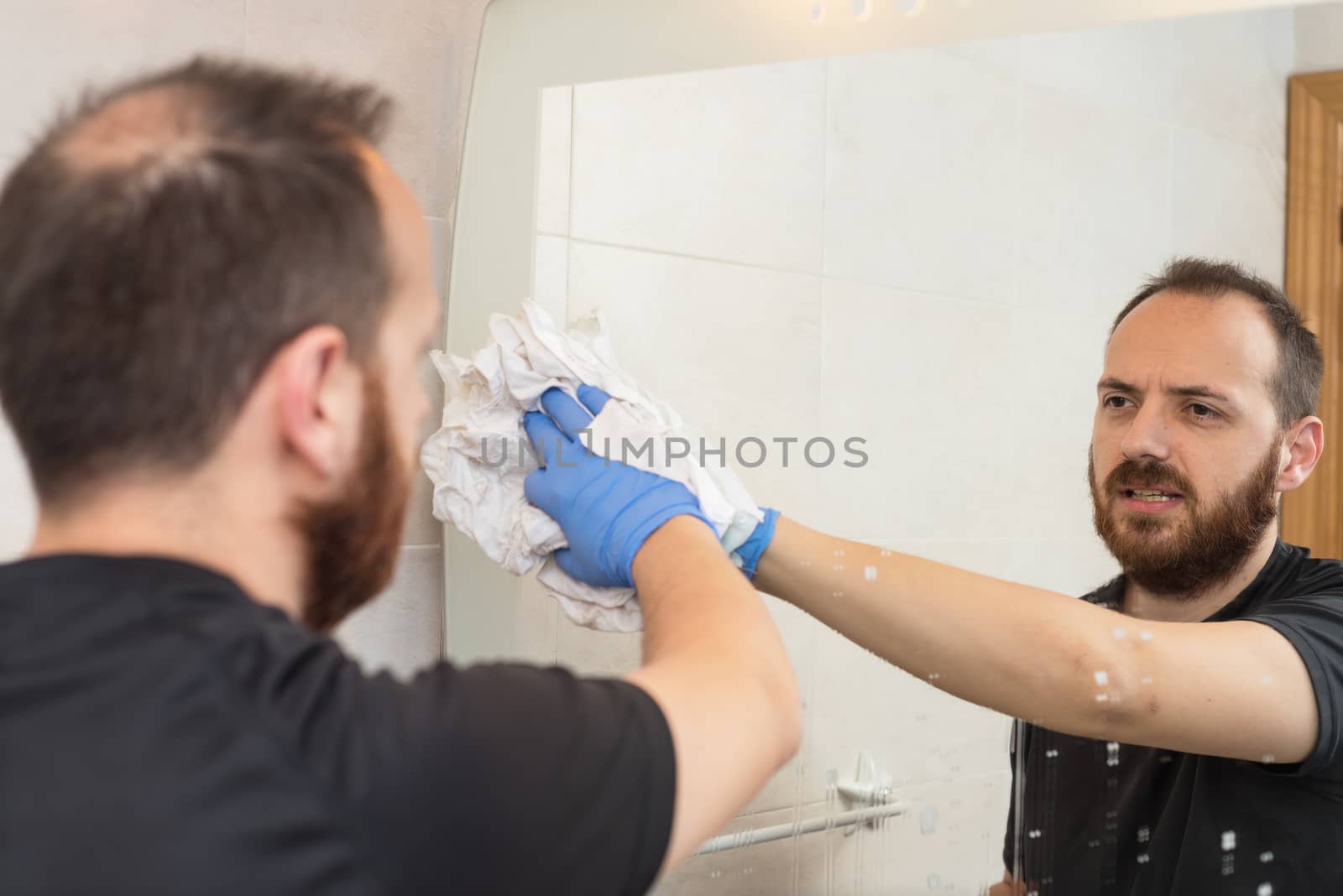 Cheerful man cleaning bathroom mirror by HERRAEZ