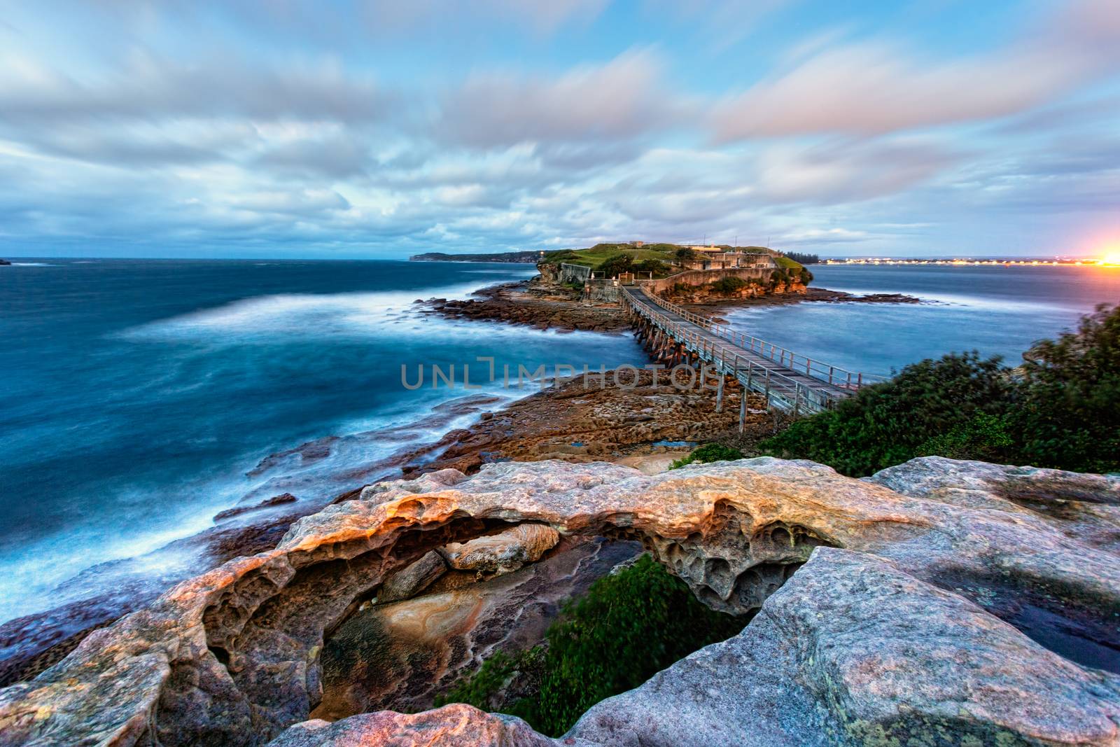 Strong winds whip up the ocean and move the clouds briskly across the sky.  A long exposure exaggerates this movement.  Views across the old timber bridge to Bare Island in the dawn hours 