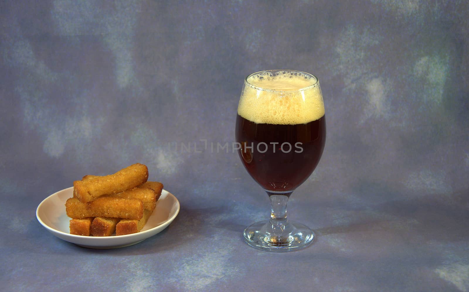 A full glass with dark beer and foam and wheat croutons on a plate. Close-up.