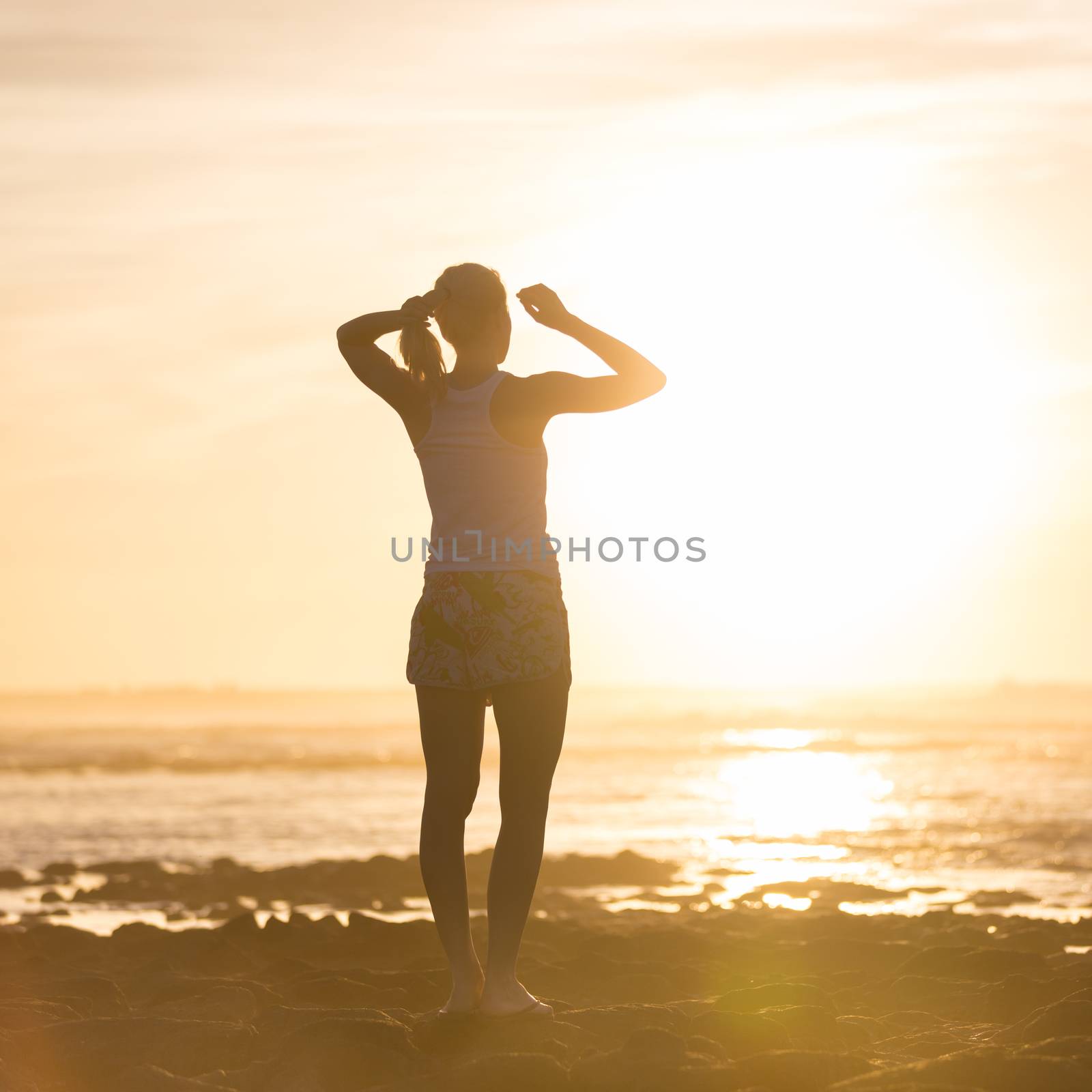 Silhouette of sporty woman clamping her hair in a ponytail while watching sunset at the beach.
