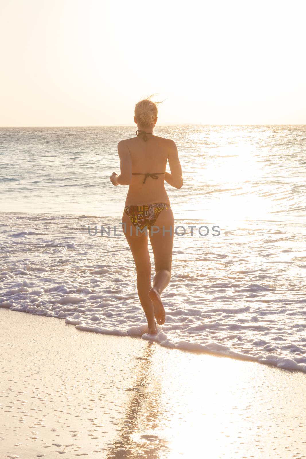 Happy woman enjoying in summer, running joyfully on tropical beach in sunset. Beautiful caucasian model wearing bikini on vacations on sandy beach. Footprints in sand.