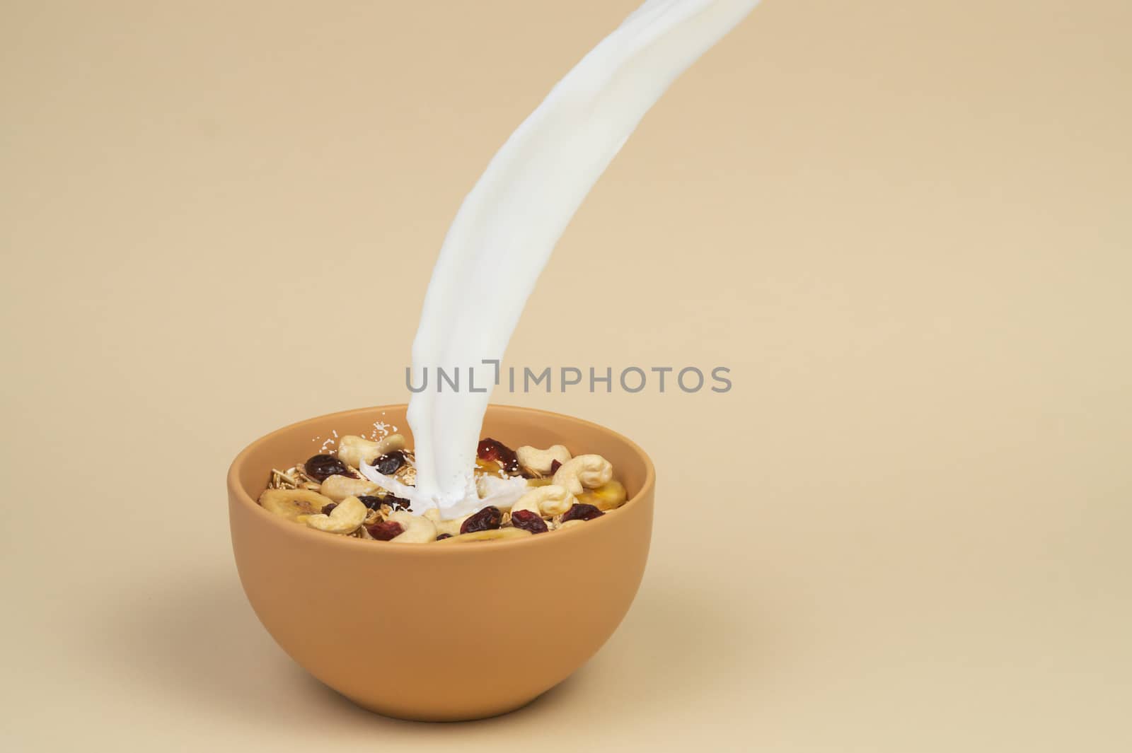 A stream og milk pours onto granola with dried and candied fruits and nuts in beige colour bowl. Bowl on a beige background. Concept of nutrient and healty breakfast or meal