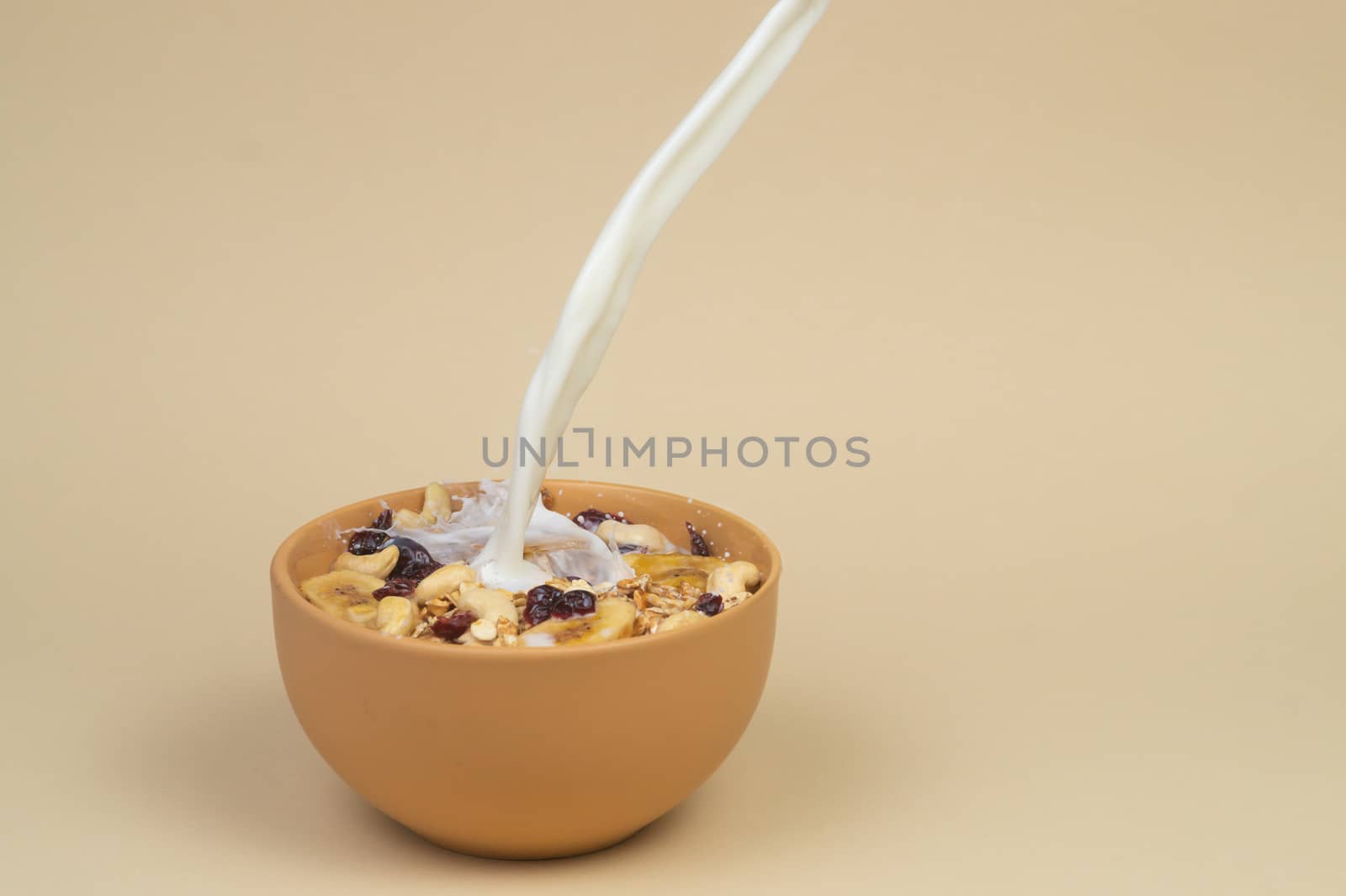 A stream og milk pours onto granola with dried and candied fruits and nuts in beige colour bowl. Bowl on a beige background. Concept of nutrient and healty breakfast or meal