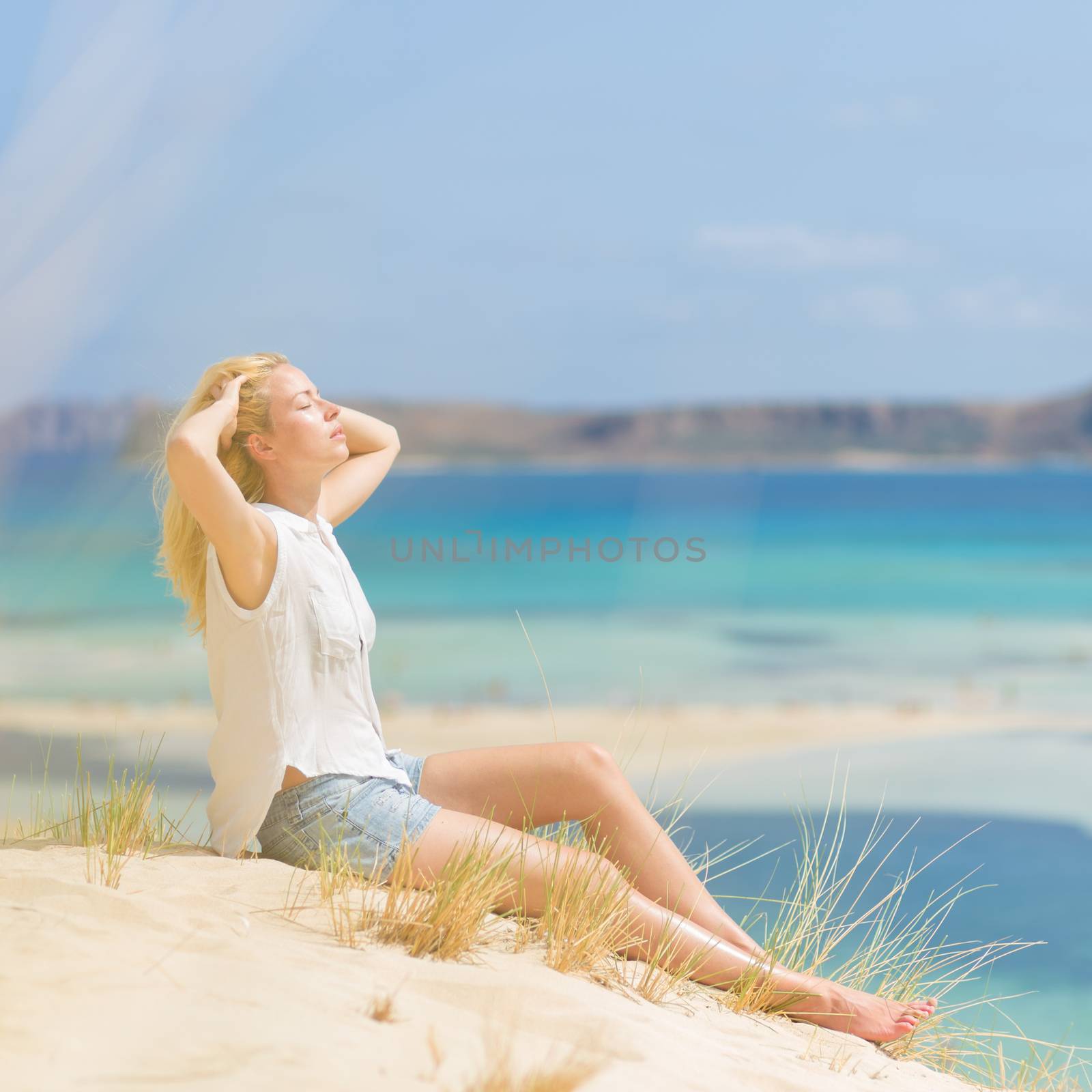 Relaxed woman enjoying sun, freedom and life an a beautiful sandy beach of Balos in Greece. Young lady feeling free, relaxed and happy. Vacations, freedom, happiness, enjoyment and well being.