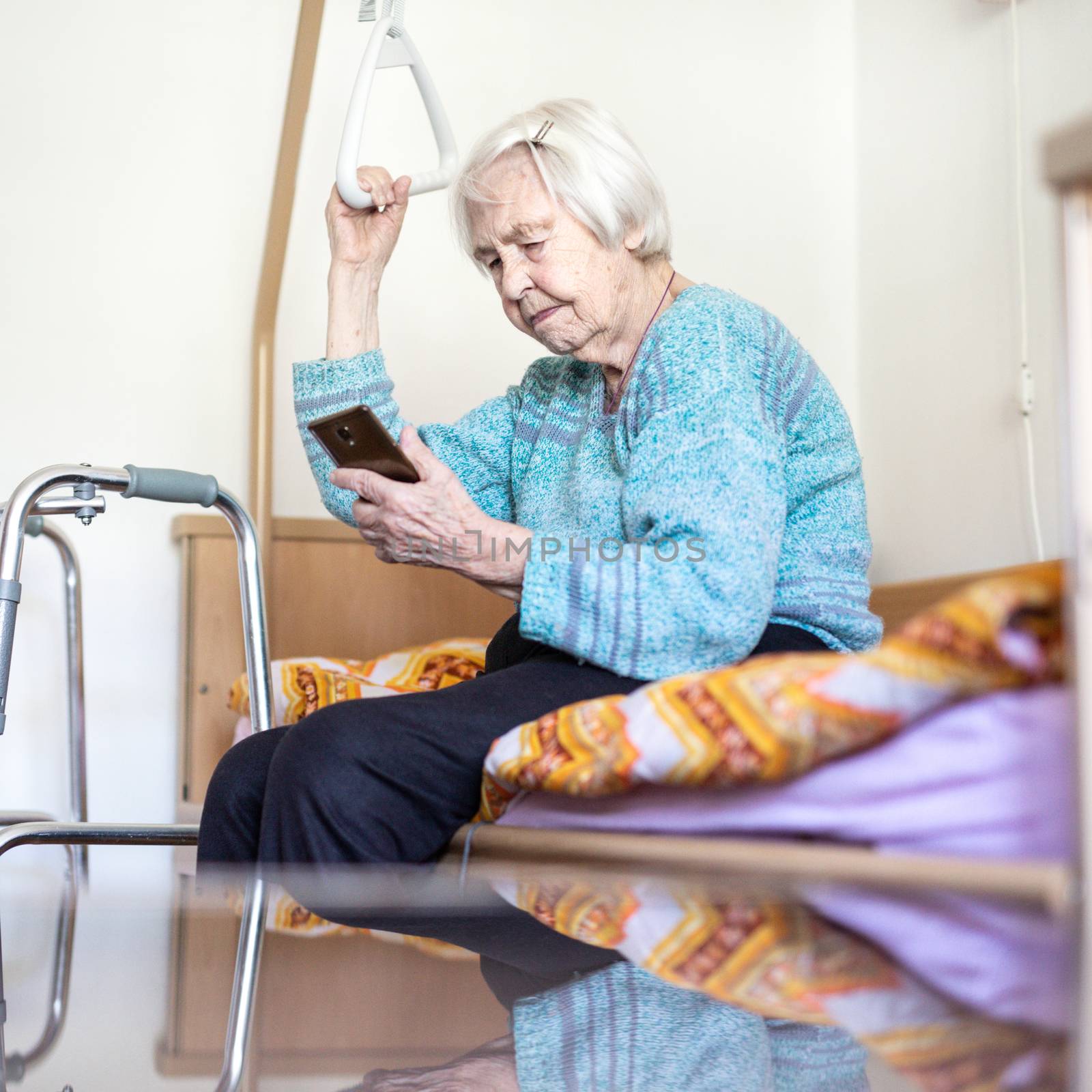 Elderly 96 years old woman reading phone message while sitting on medical bed supporting her by holder. Senior woman using wireless internet connection on smart phone.