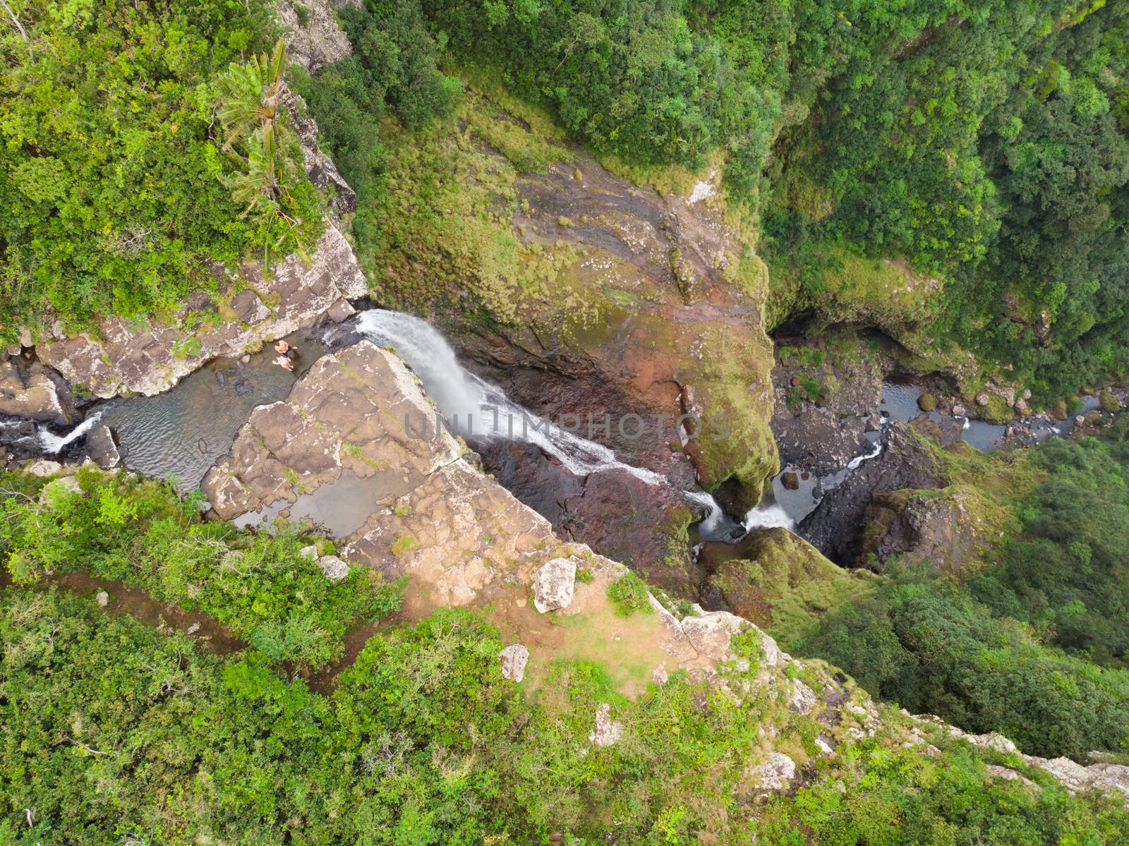 Aerial top view of travel couple waving to drone, standing on the edge of 500 feet waterfall in the tropical island jungle of Black river gorges national park on Mauritius island.