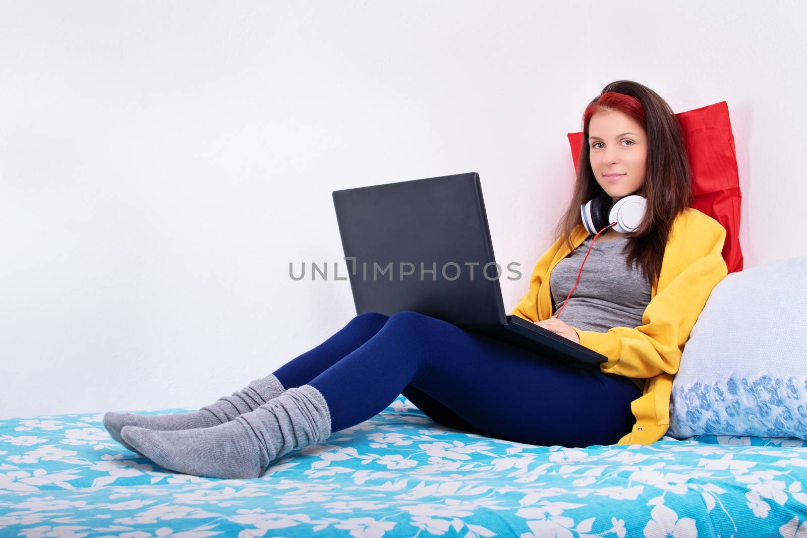 Beautiful smiling young girl sitting on her bed with a laptop in her lap and headphones around her neck.