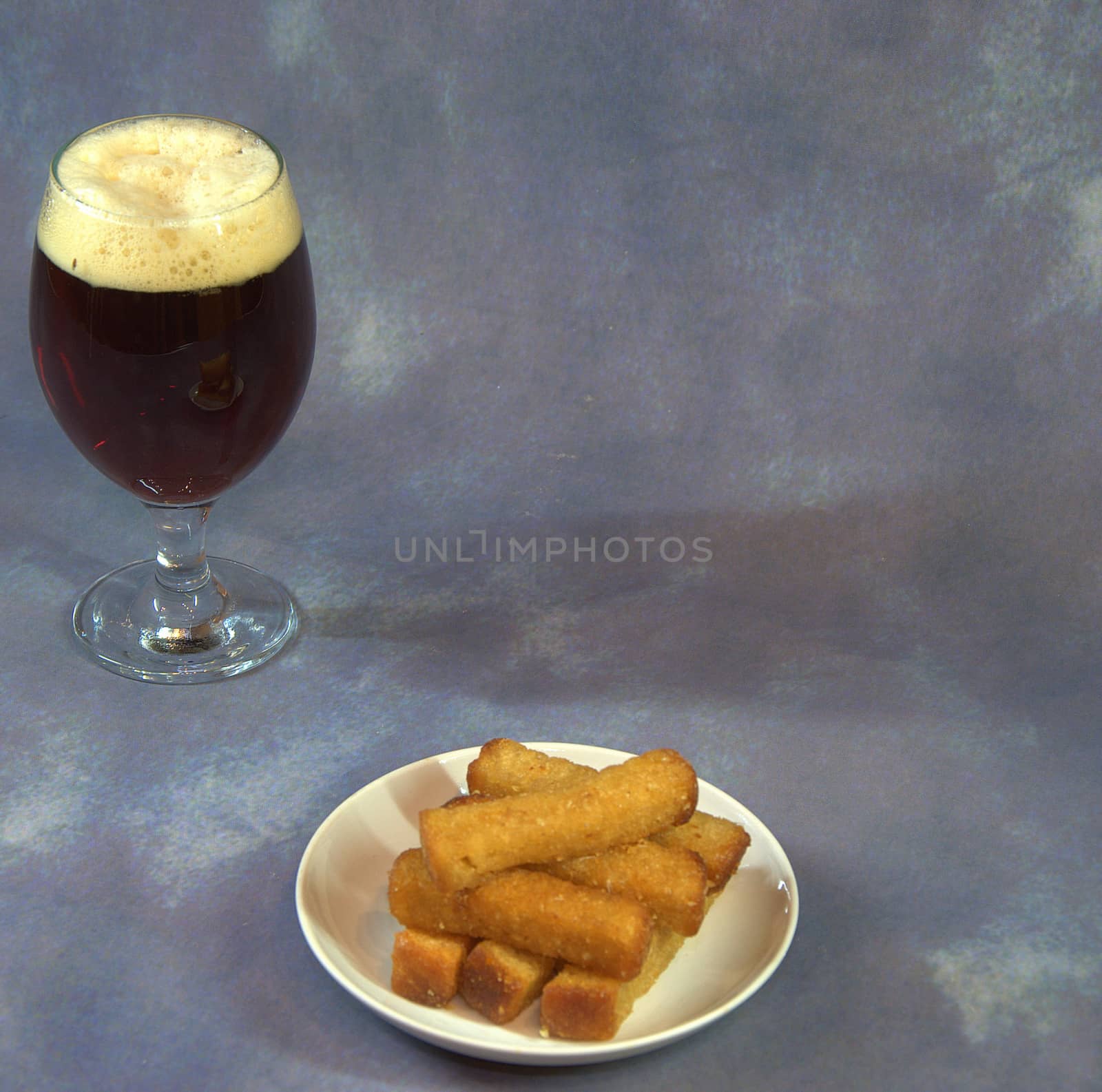 A full glass of dark beer with foam and a plate with wheat croutons on a gray background. Close-up.