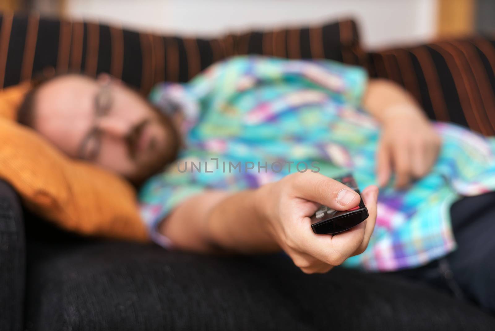 Man relaxing in sofa with remote control watching tv. Shallow dof focus on remote control. by HERRAEZ