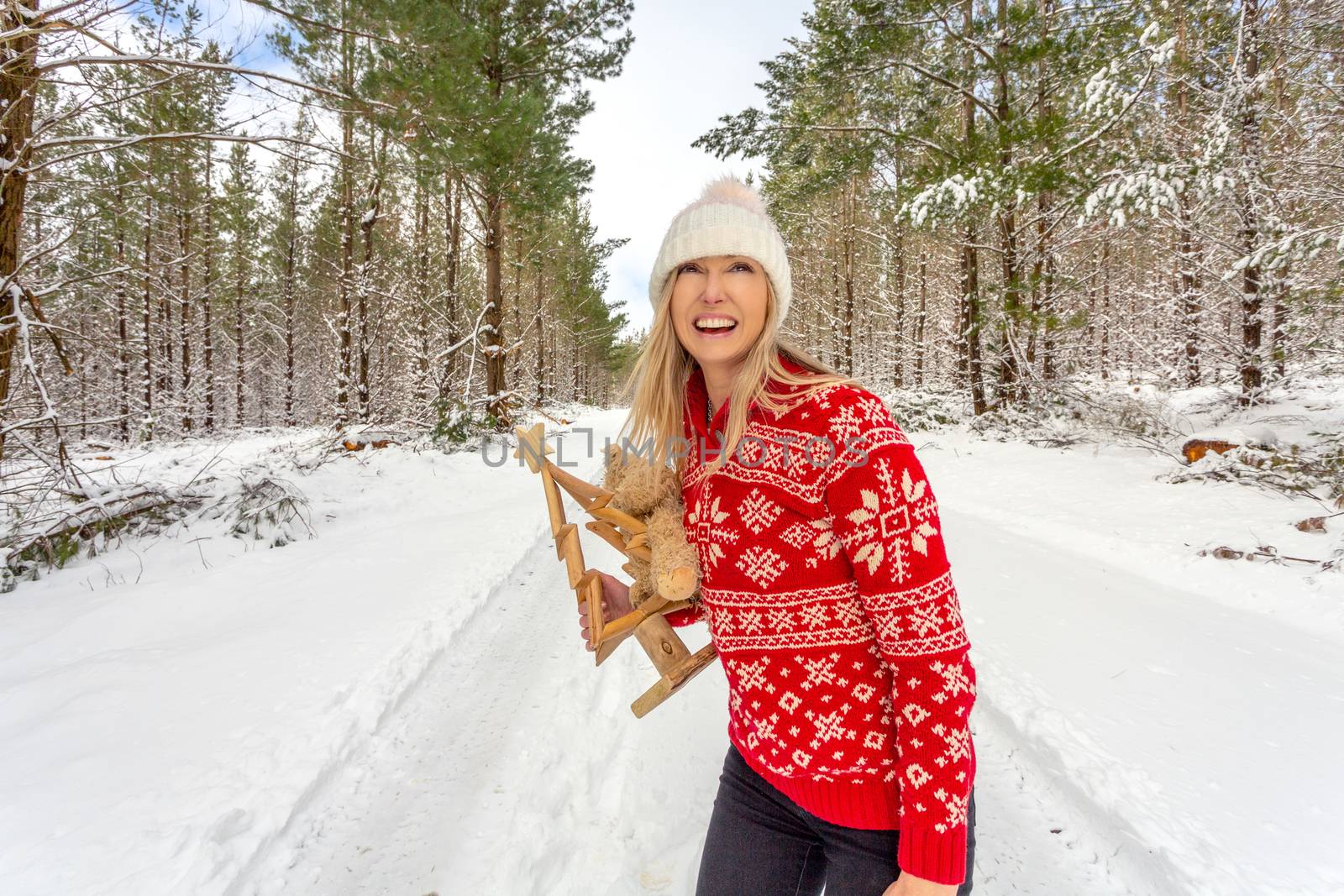 Happy cheerful woman on a snow covered road at Christmas time through tall pine trees covered in snow.  She has a cheerful expression and wearing a red jumper with snowflakes on it.