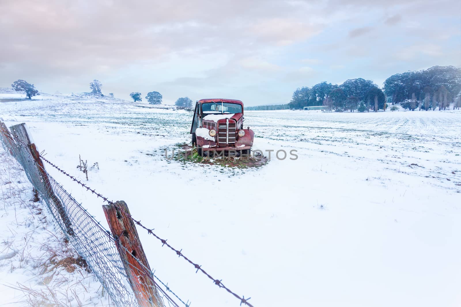 A rusty old vintage car sits in a snow covered field in winter.   There is a leaning rusty barbed wire fence in the foreground .   Space for copy