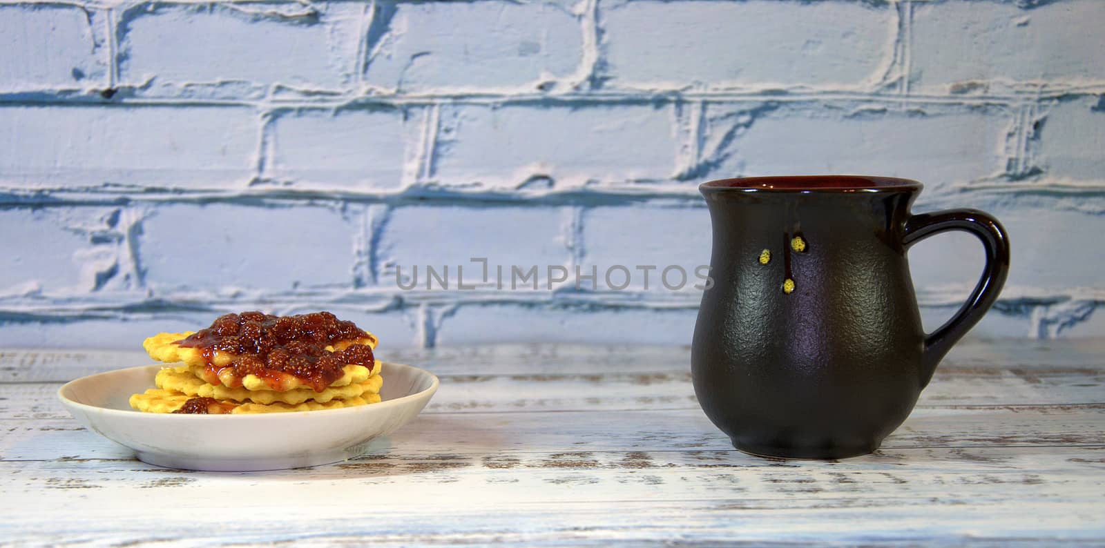 Dark ceramic cup with tea, next to a plate with waffles with strawberry jam. Close-up.