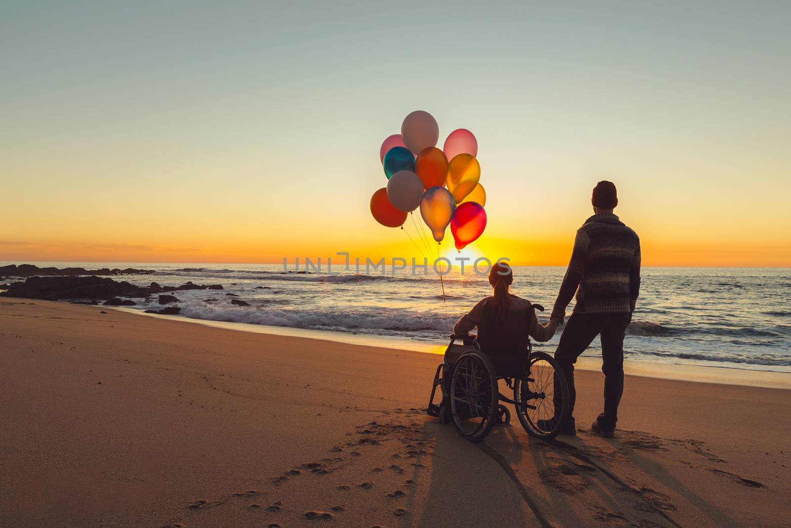 Happy couple at the beach, where woman is on a wheelchair holding balloons on her hands