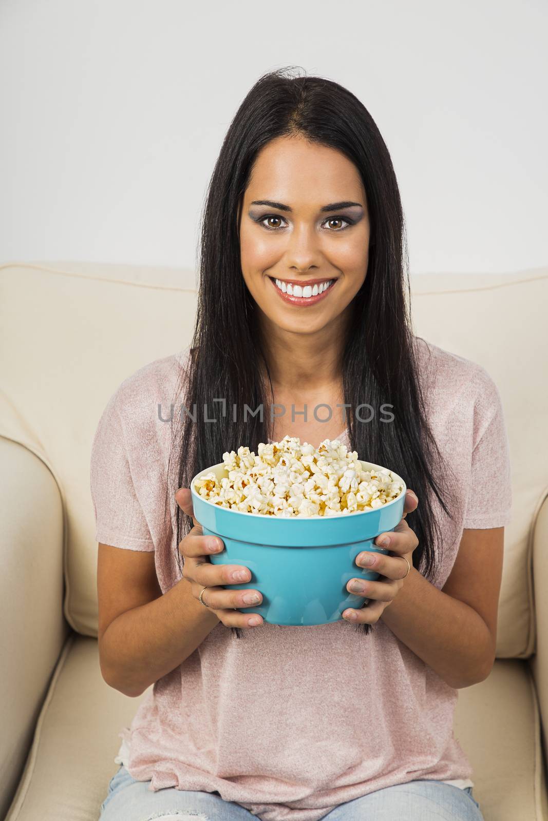 Happy young woman, sitting on a white couch, holding a blue bowl of popcorn
