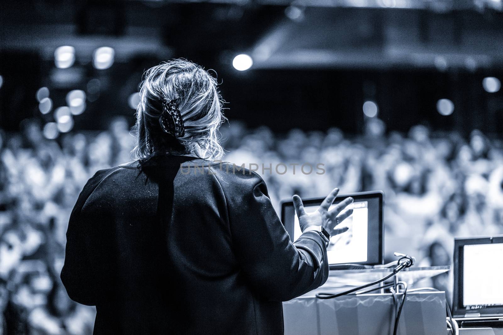 Female speaker giving a talk on corporate business conference. Unrecognizable people in audience at conference hall. Business and Entrepreneurship event. Black and white, blue toned image.