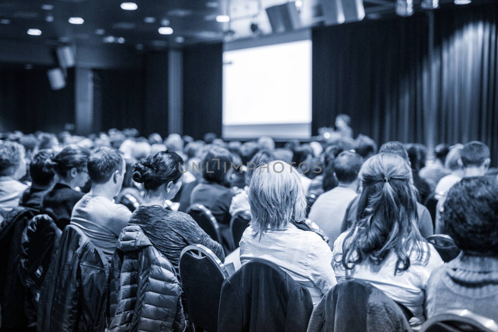 Business and entrepreneurship symposium. Speaker giving a talk at business meeting. Audience in conference hall. Rear view of unrecognized participant in audience.