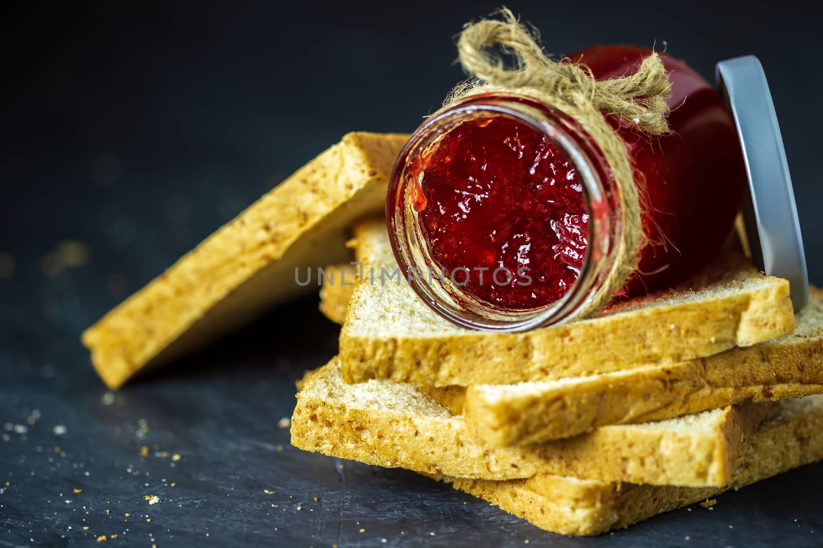 Strawberry jam bottle and whole wheat bread are stacked on a black background. Concept of breakfast and healthy food.