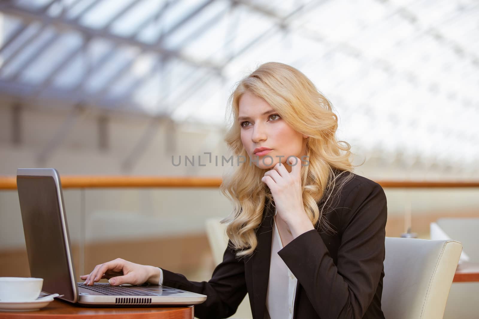 Portrait of young pretty blonde woman sitting in cafe with her laptop, typing and thinking