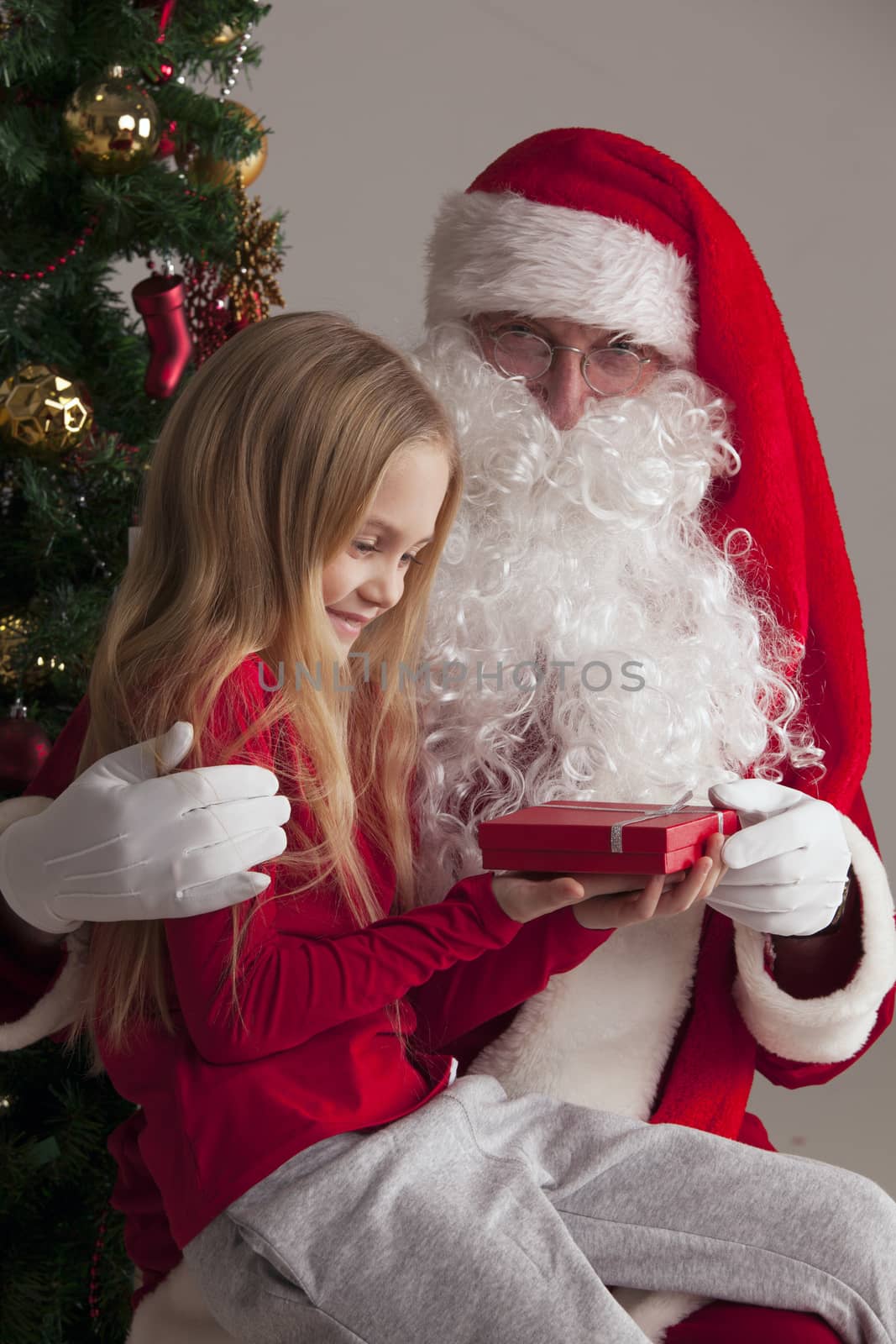 Portrait of smiling little girl sitting on santa claus knees near christmas tree and getting a gift