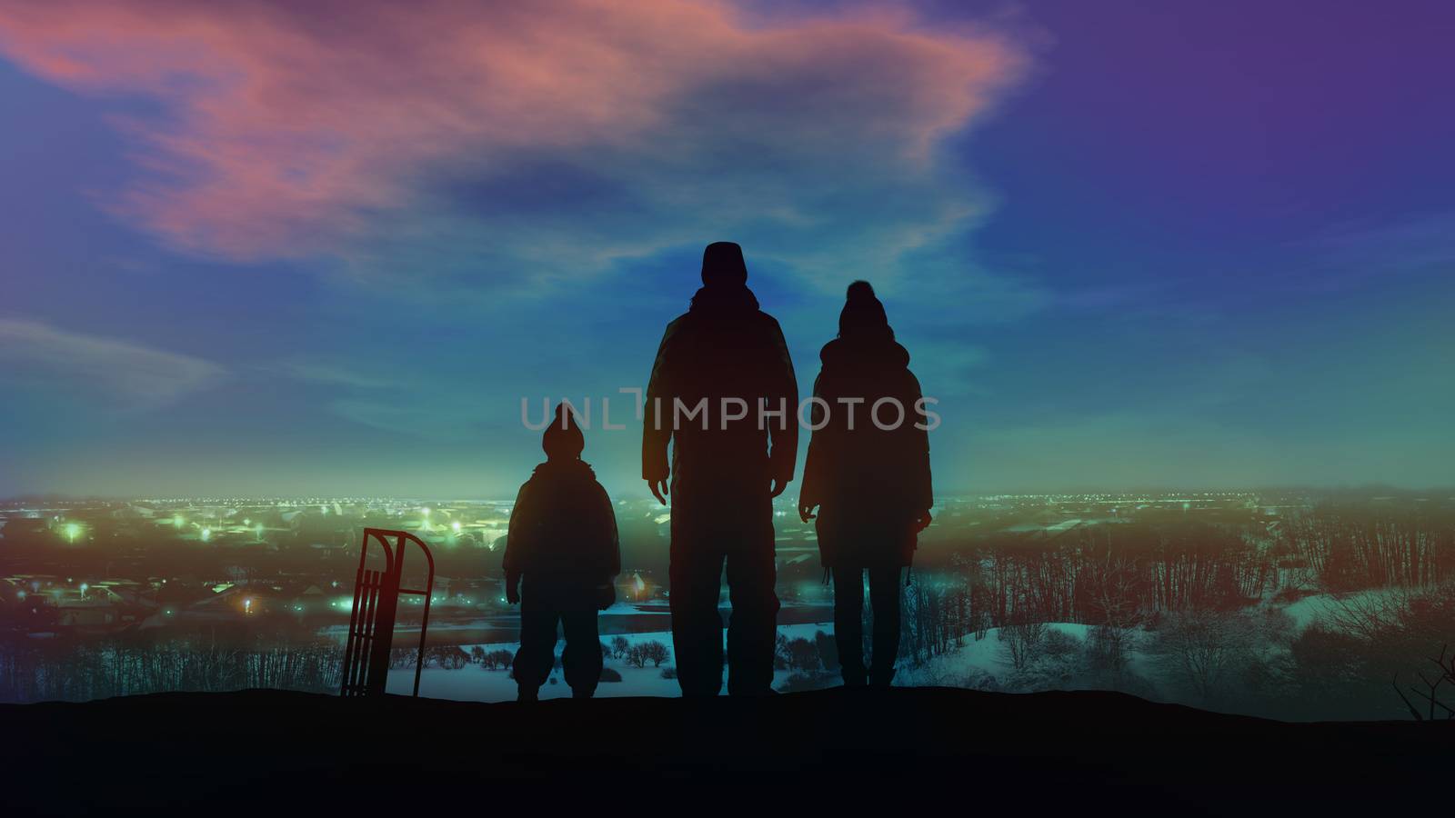 Family of three standing on a hill look at the lights of the Christmas village.