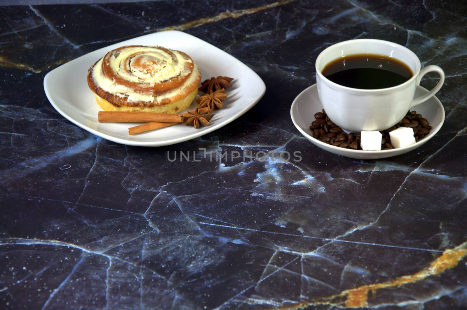A cup of black coffee with two pieces of sugar in a saucer and a plate with a bun in the glaze with cinnamon sticks and star anise. Close-up.