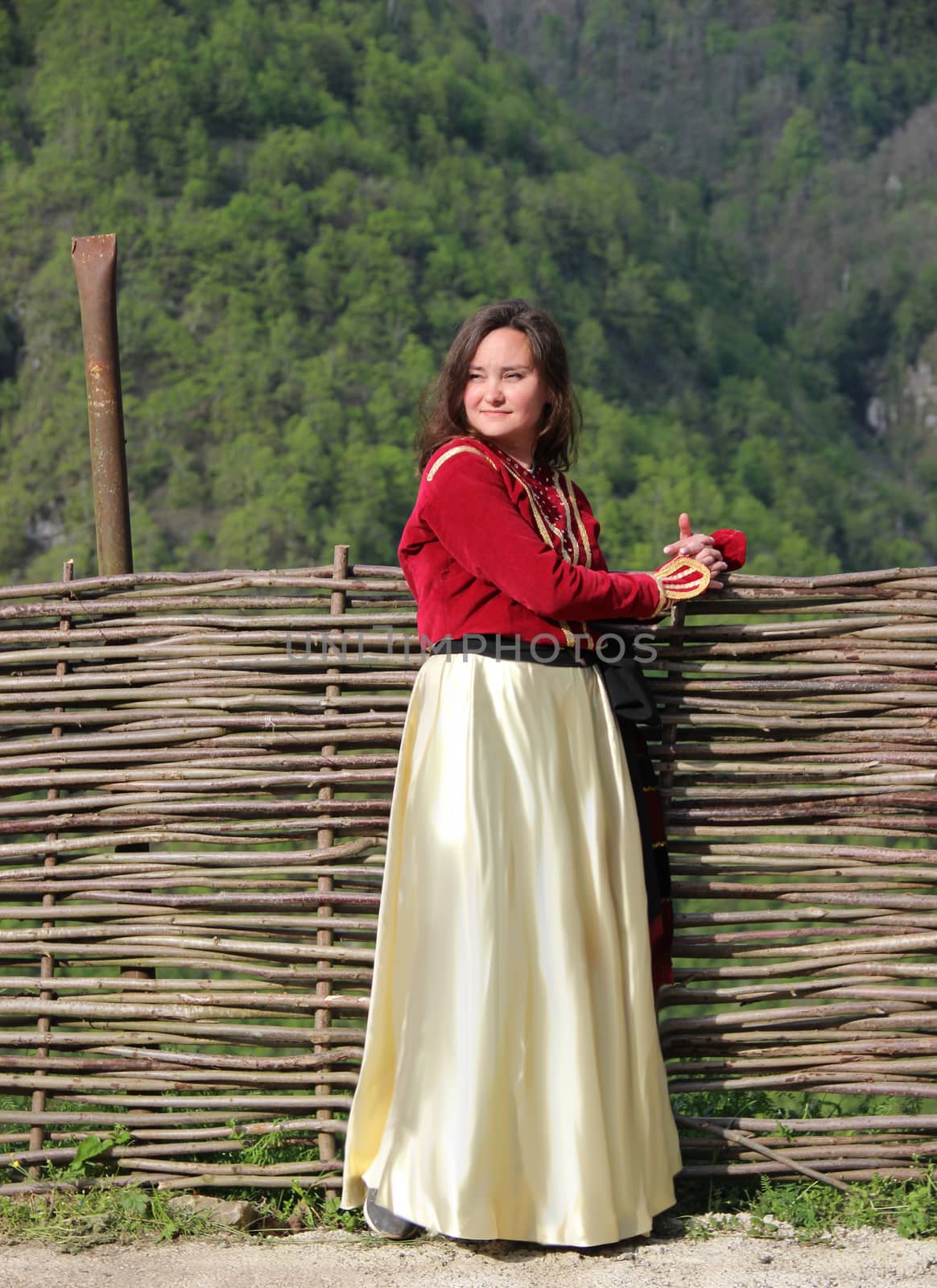 Photo of young brunette woman in a traditional georgian costume stands near a wicker fence on a background of mounains covered in a freen forest