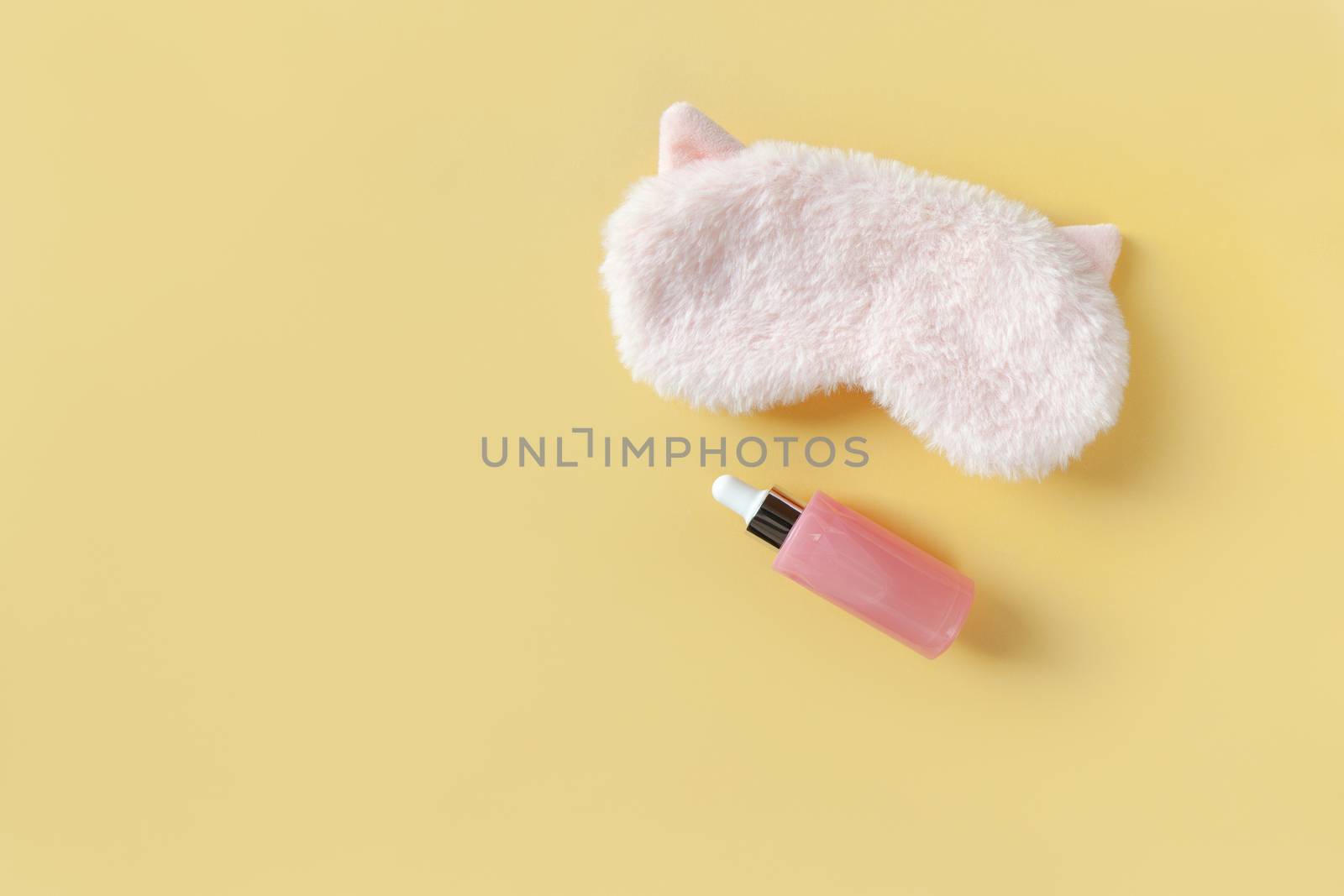 Pink fluffy fur sleep mask with small ears and bottle with serum for face on pastel yellow background. Top view. Concept of vivid dreams and skin care. Minimal style, copy space. Horizontal by ALLUNEED