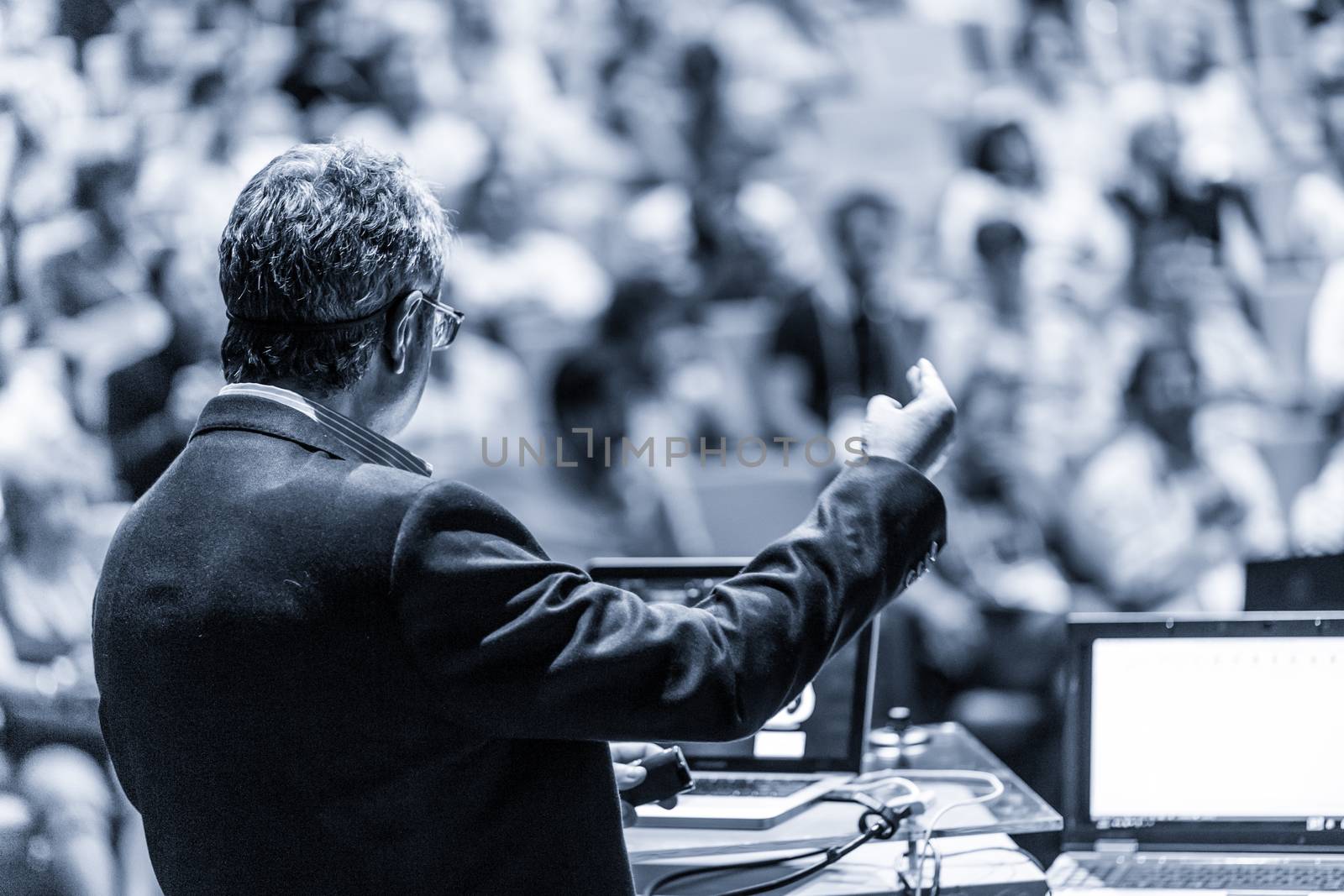 Speaker giving a talk on corporate business conference. Unrecognizable people in audience at conference hall. Business and Entrepreneurship event. Black and white, blue toned image.