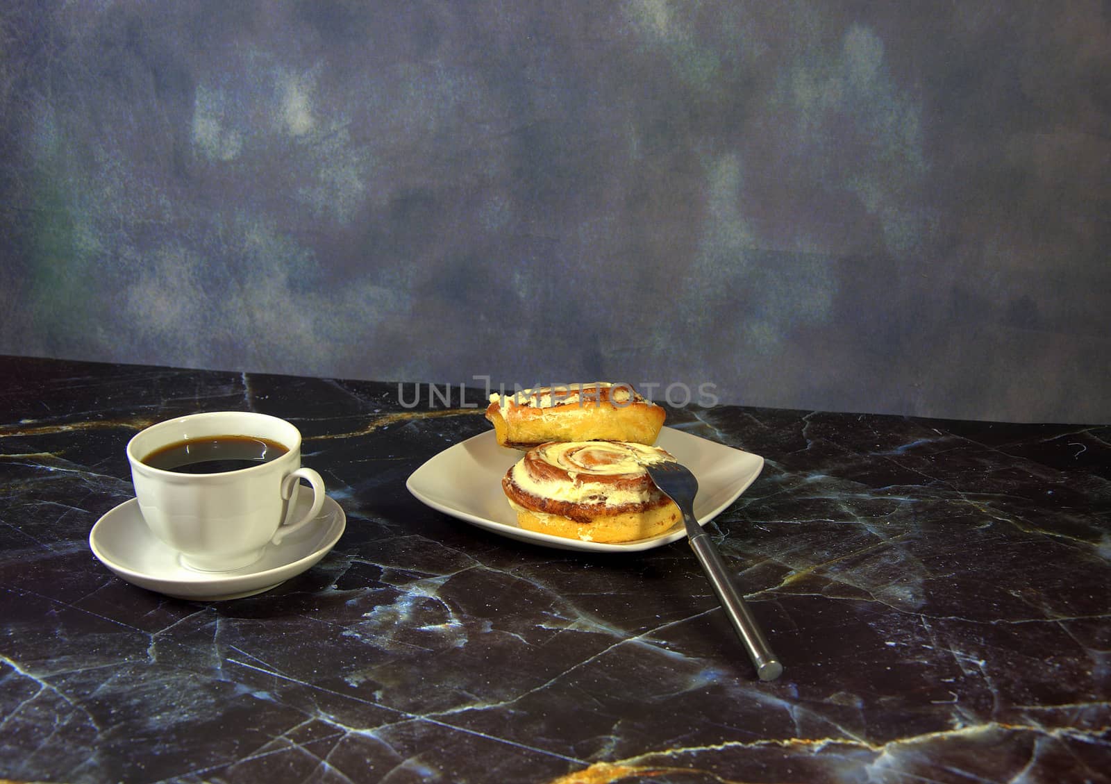 A cup of black coffee on a saucer and bwe cinnamon rolls on a plate, next to a fork. Close-up.
