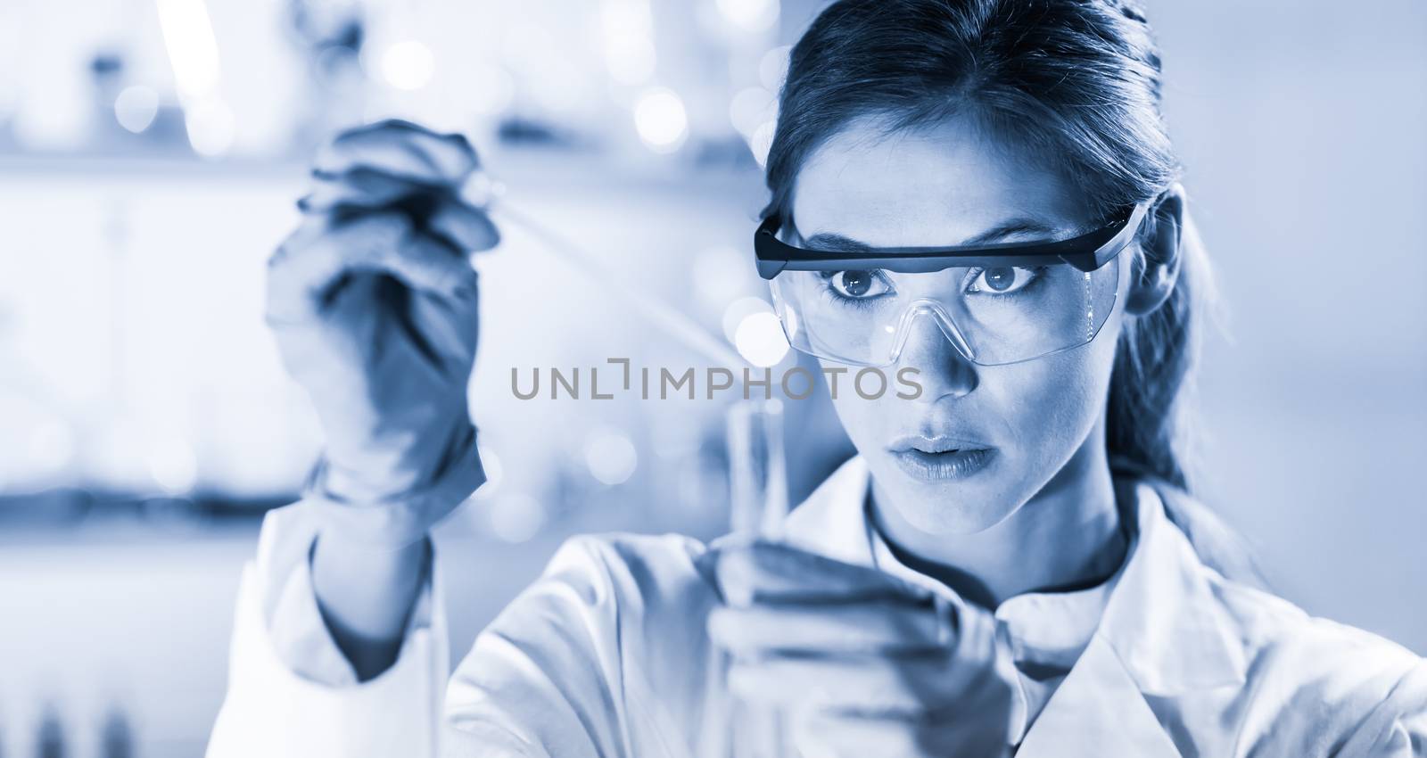 Life scientists researching in laboratory. Focused female life science professional pipetting solution into the glass cuvette. Healthcare and biotechnology concept. Blue toned image.