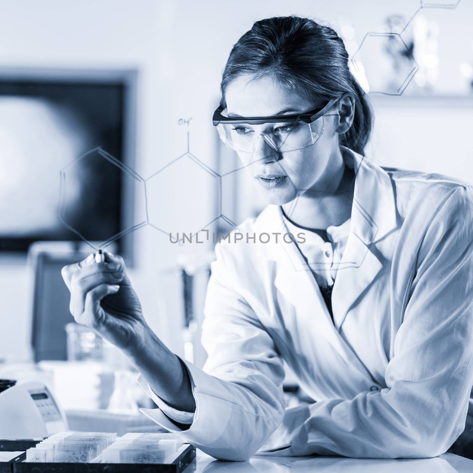Portrait of a confident female health care expert in life science laboratory writing structural chemical formula on a glass board. Healthcare and modern life science concept. Blue toned image.