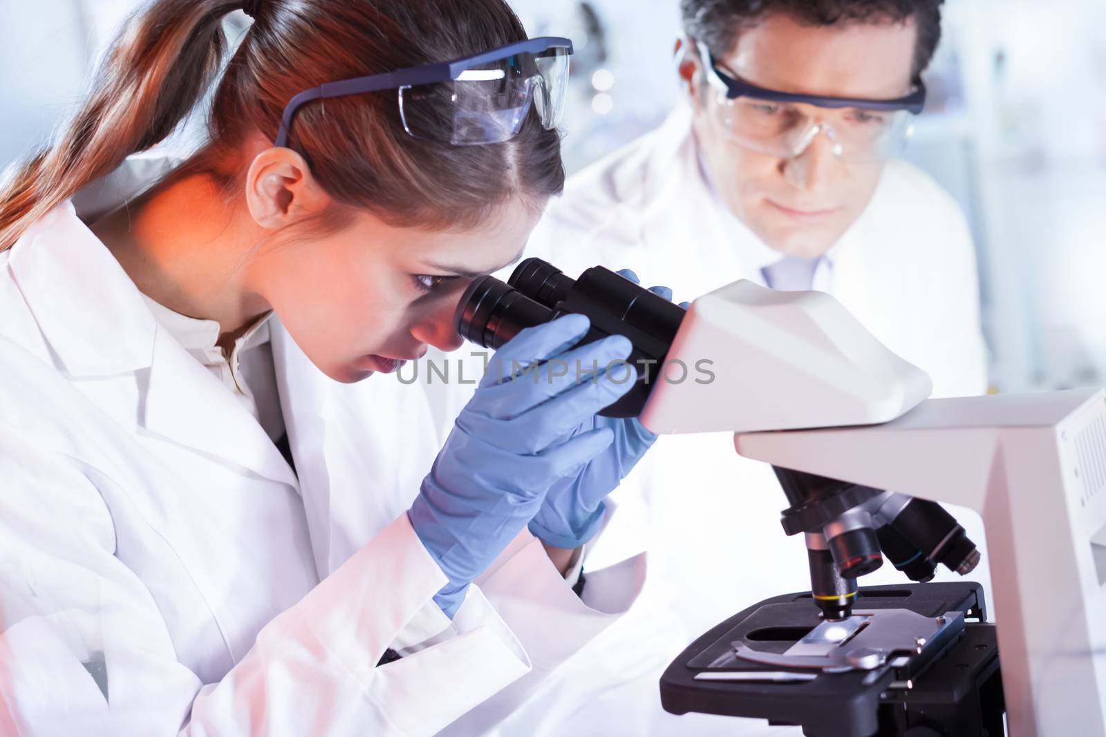 Life scientists researching in laboratory. Attractive female young scientist and her doctoral supervisor microscoping in their working environment. Healthcare and biotechnology.