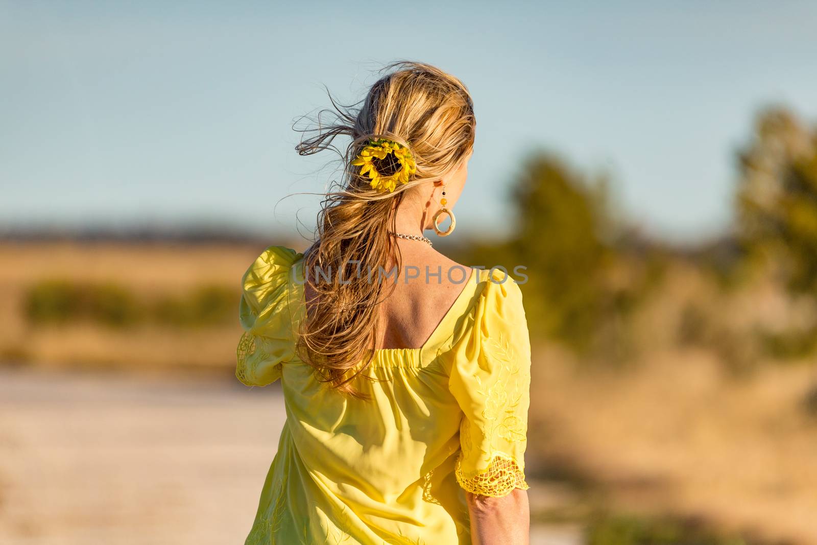 Woman outdoors in beautiful sunshine rural area.  She has a sunflower in her long wavy hair blowing gently in the wind.