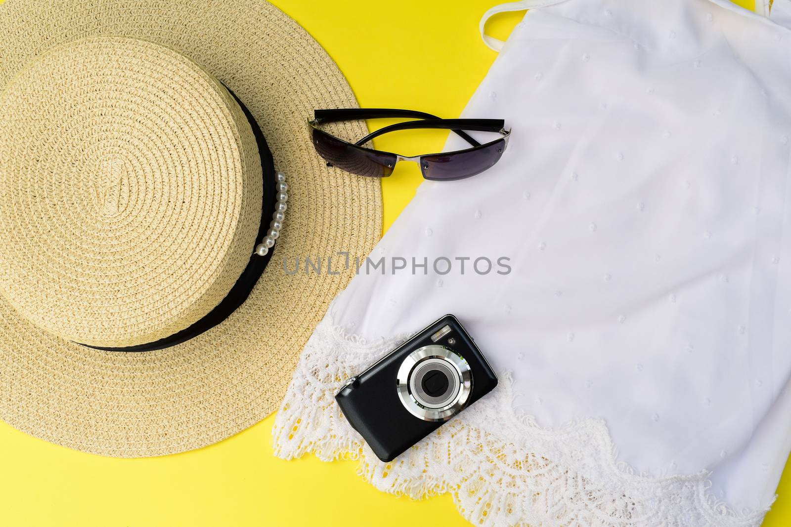 Summer travel concept on yellow background. Straw hat with beaded ribbon decoration. Black photo camera and sunglasses on white lacy female top.