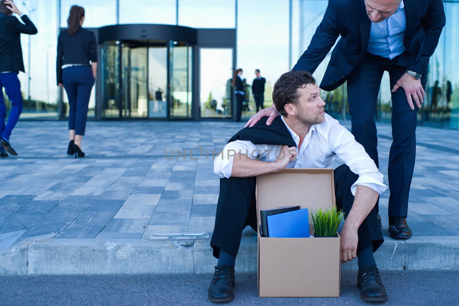 Fired business man sitting frustrated and upset on the street near office building with box of his belongings. He lost work. Business man comforts and encourages him