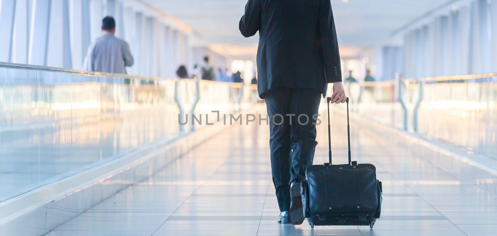 Businessman walking and wheeling a trolley suitcase at the lobby, talking on a mobile phone. Business travel concept. by kasto