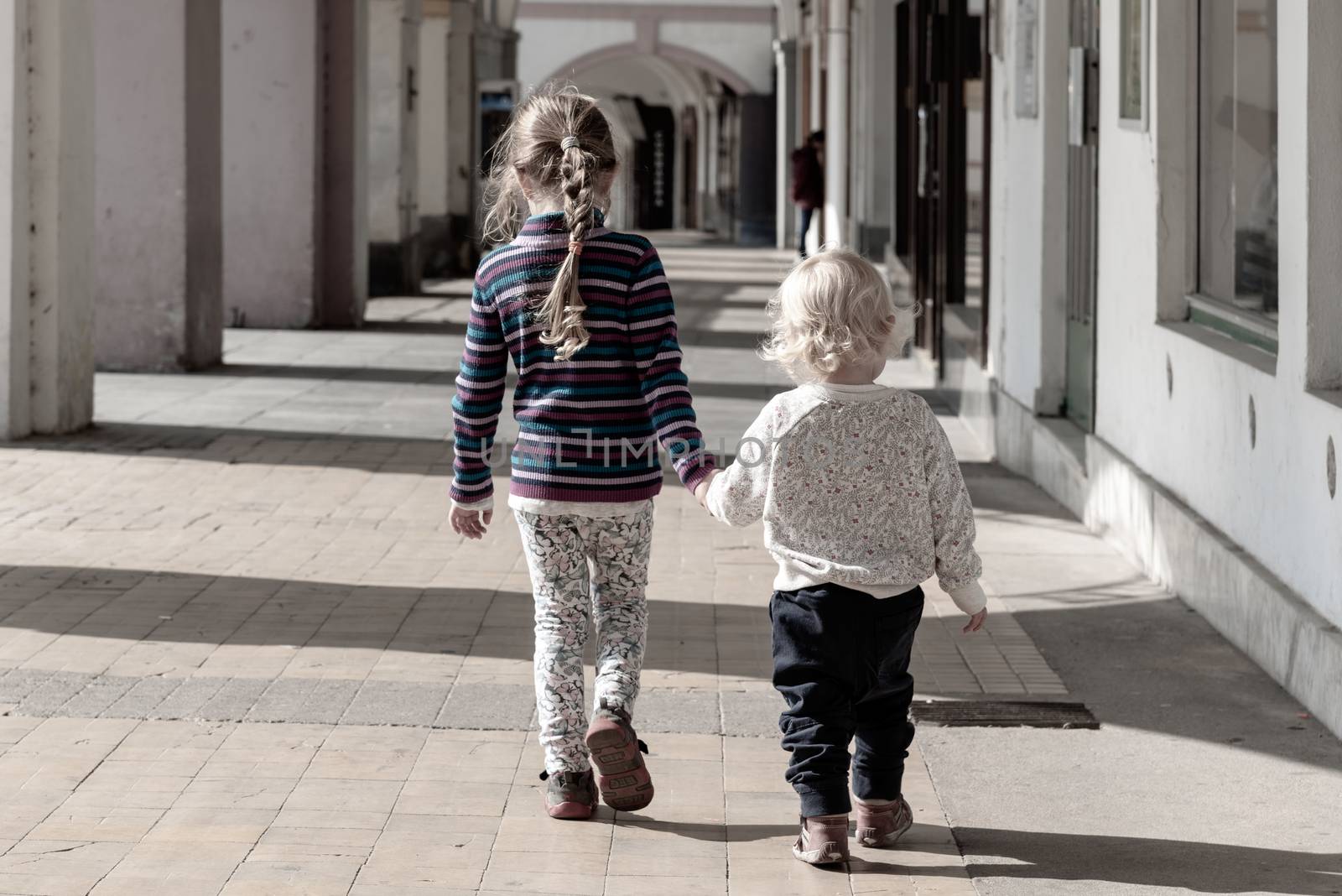 Two young children walk together hand in hand in the old town arcade street. Family love and friendship theme by pyty