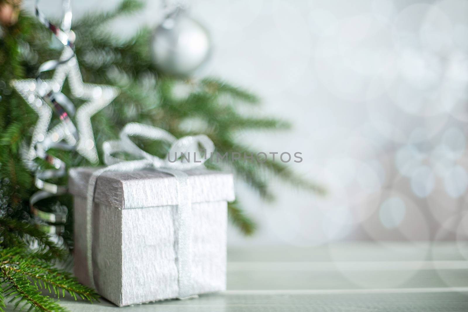 Silver Christmas new year gift with curly streamer ribbon bow over glittering bokeh lights background under fir tree with baubles