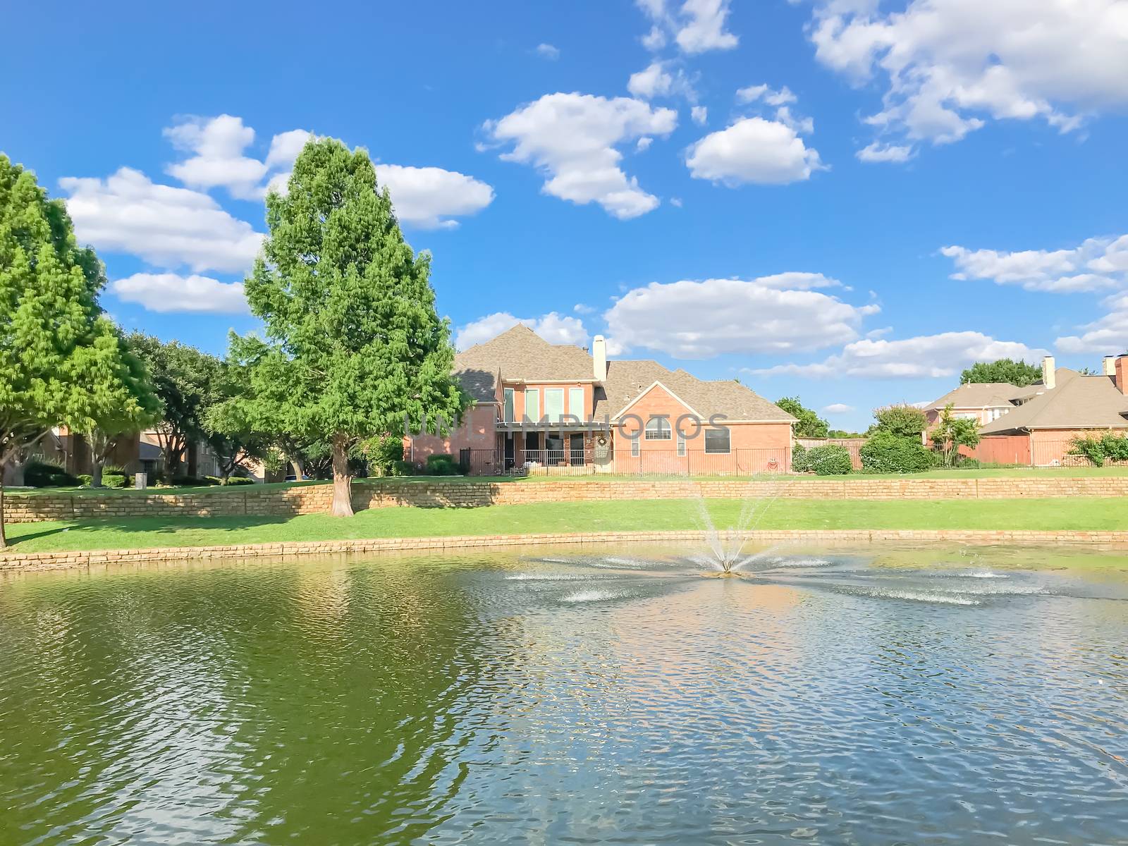 Lakefront houses with water fountain and green grass lawn bank in suburbs Dallas, Texas, USA. Suburban single family detached home along river with high stone retaining wall, sewage, mature tree