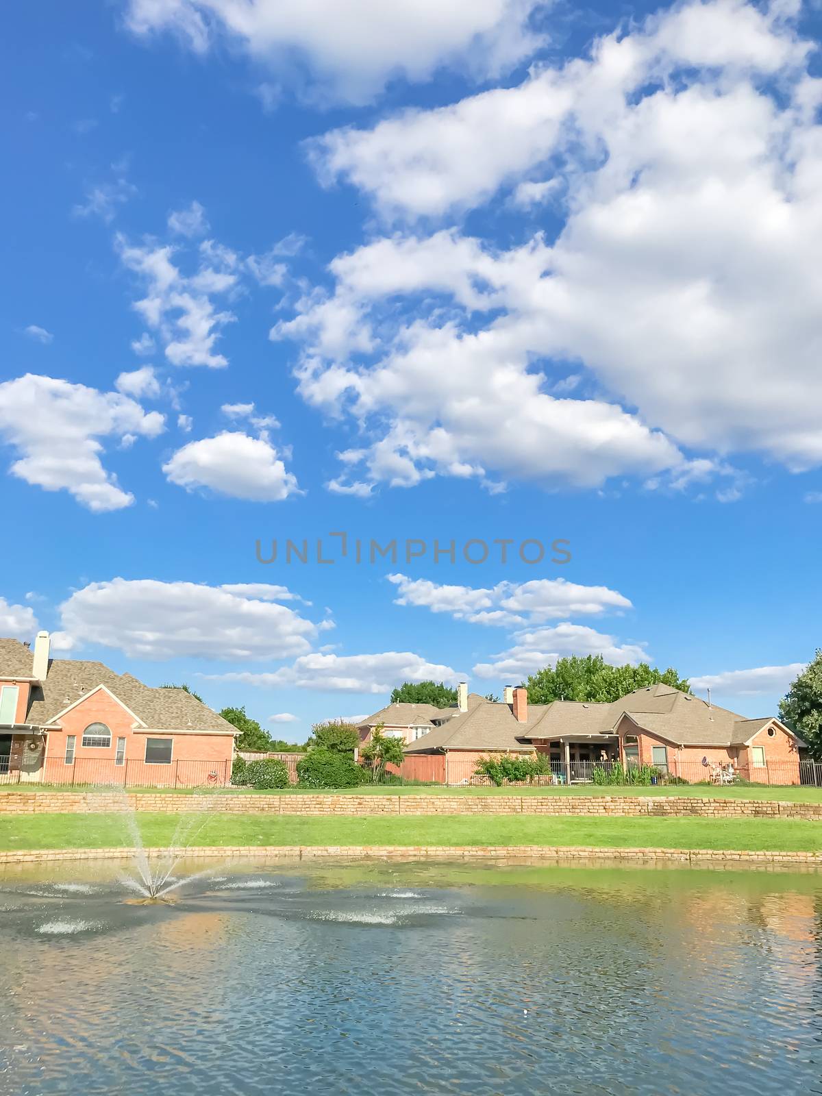 Lakefront houses with water fountain and green grass lawn bank in suburbs Dallas, Texas, USA. Suburban single family detached home along river with high stone retaining wall, sewage, mature tree