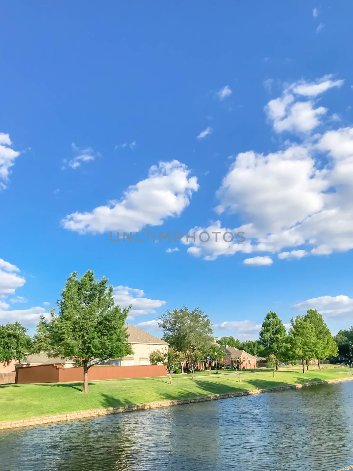 Beautiful waterfront house with row of mature bold cypress trees in suburbs Dallas, Texas, USA. Suburban single family detached home along river with high stone retaining wall, green grass lawn